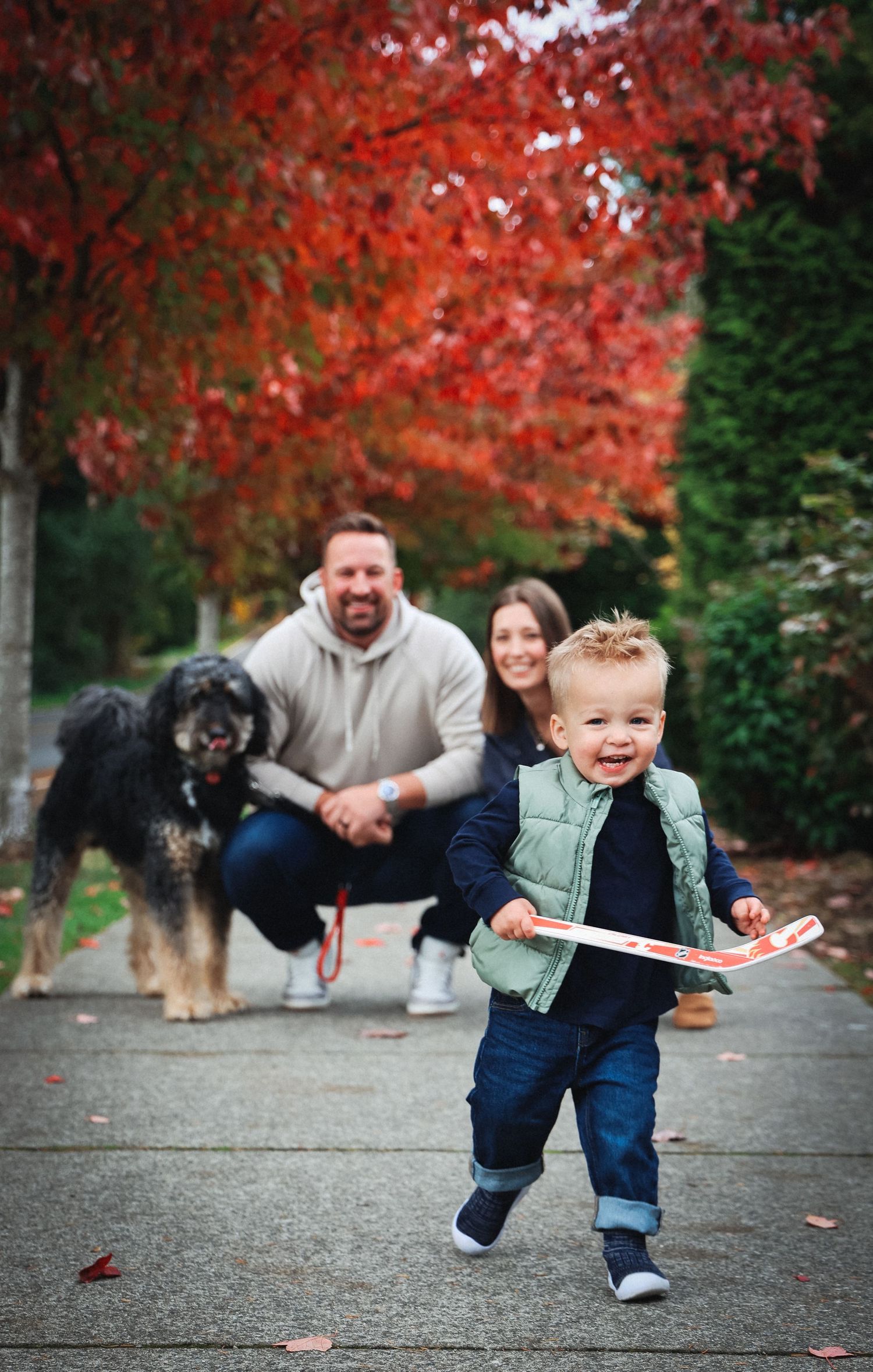Toddler running playfully under vibrant red autumn trees with family watching.