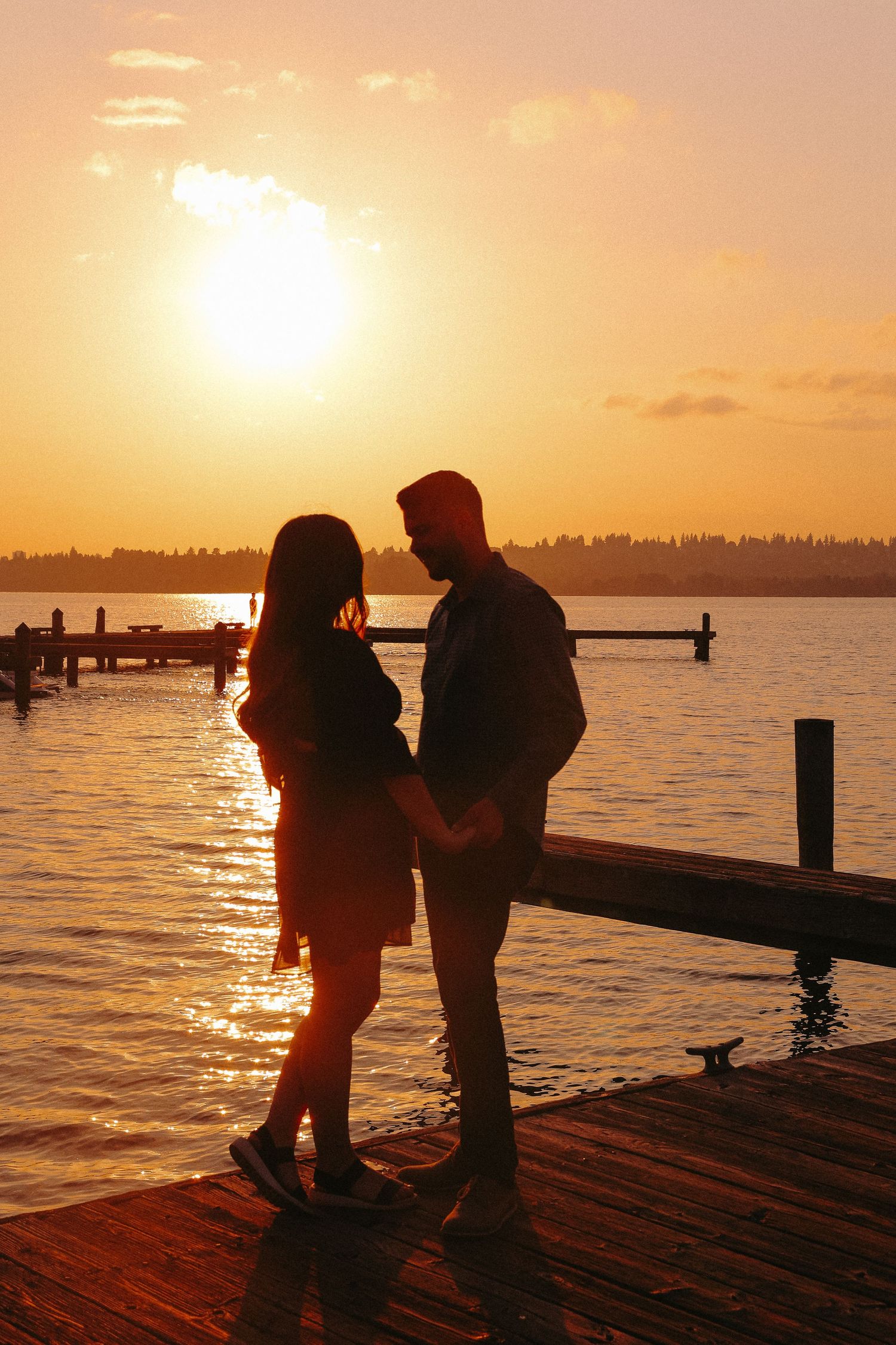 Two silhouettes stand together on a wooden dock at sunset with golden light reflecting off the calm water.