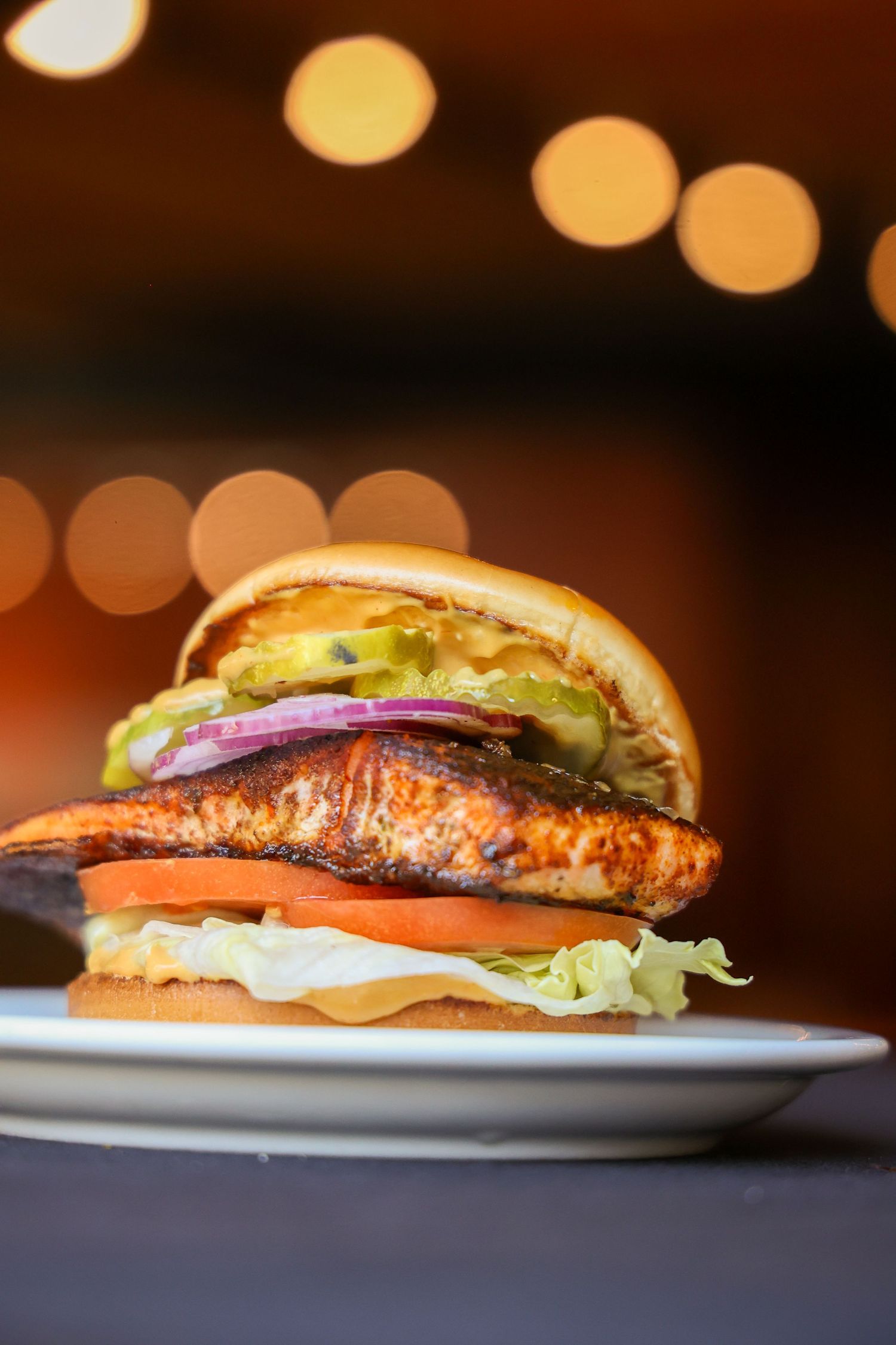 A close-up sequence of a grilled salmon burger with lettuce, tomato and red onion on a white plate against a dark background.
