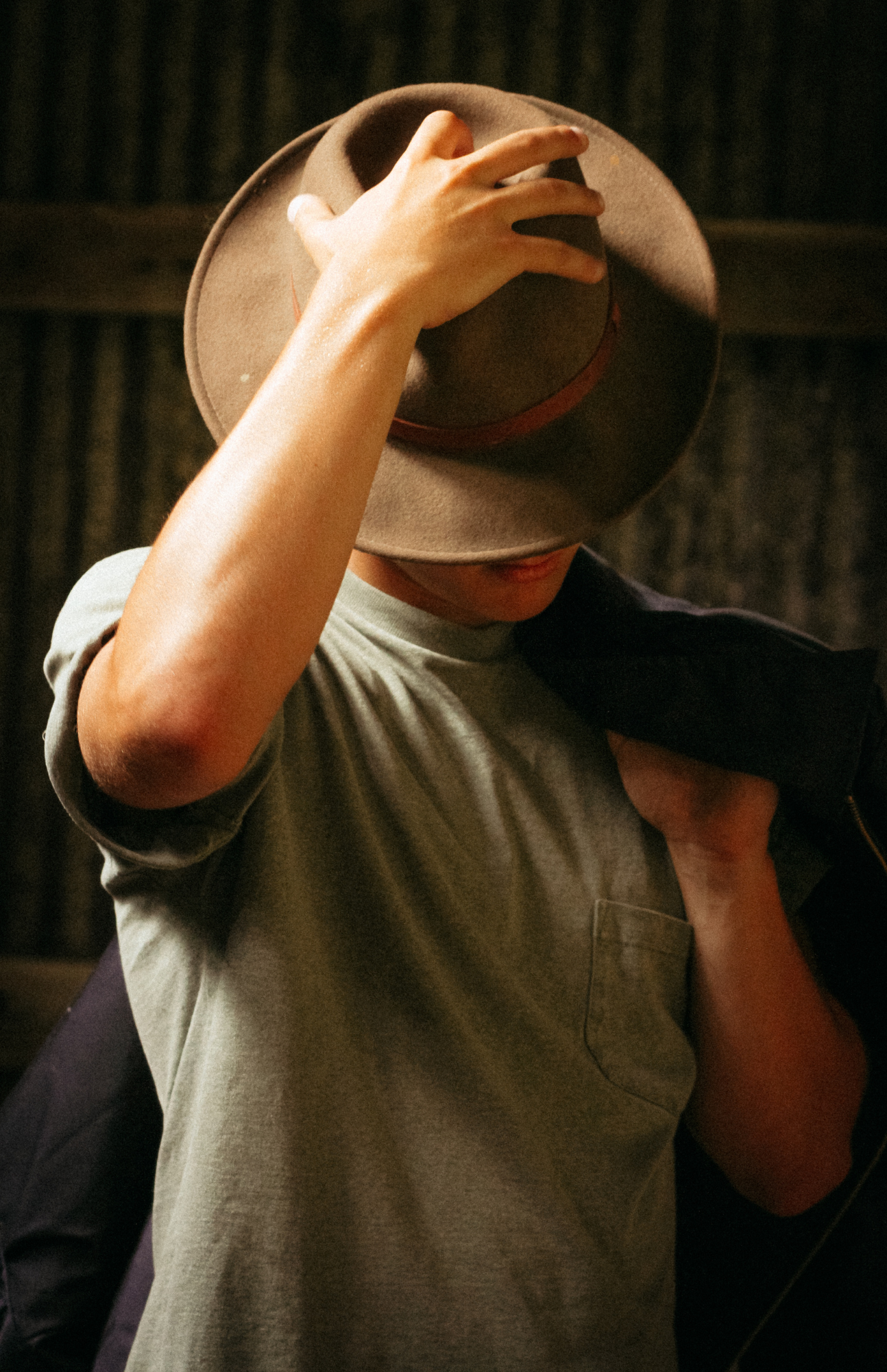Person in casual attire adjusting a brown fedora hat in dramatic lighting.