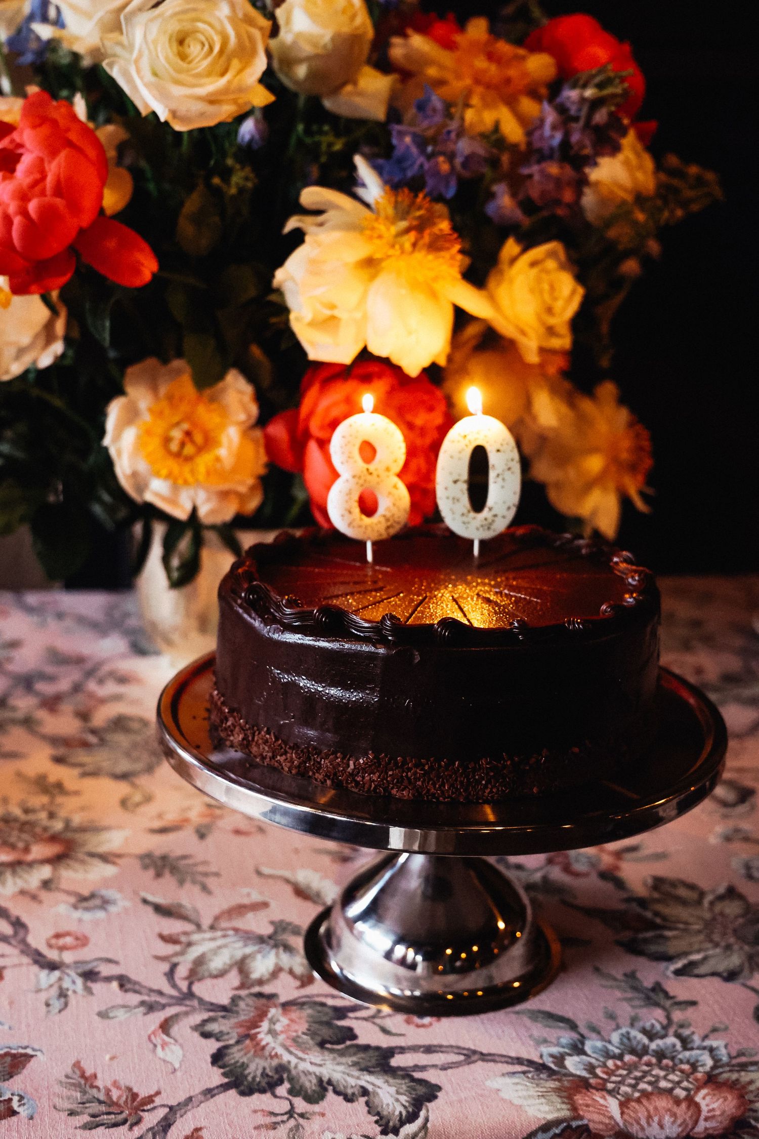 Chocolate birthday cake with number 80 candles surrounded by colorful roses on floral tablecloth.