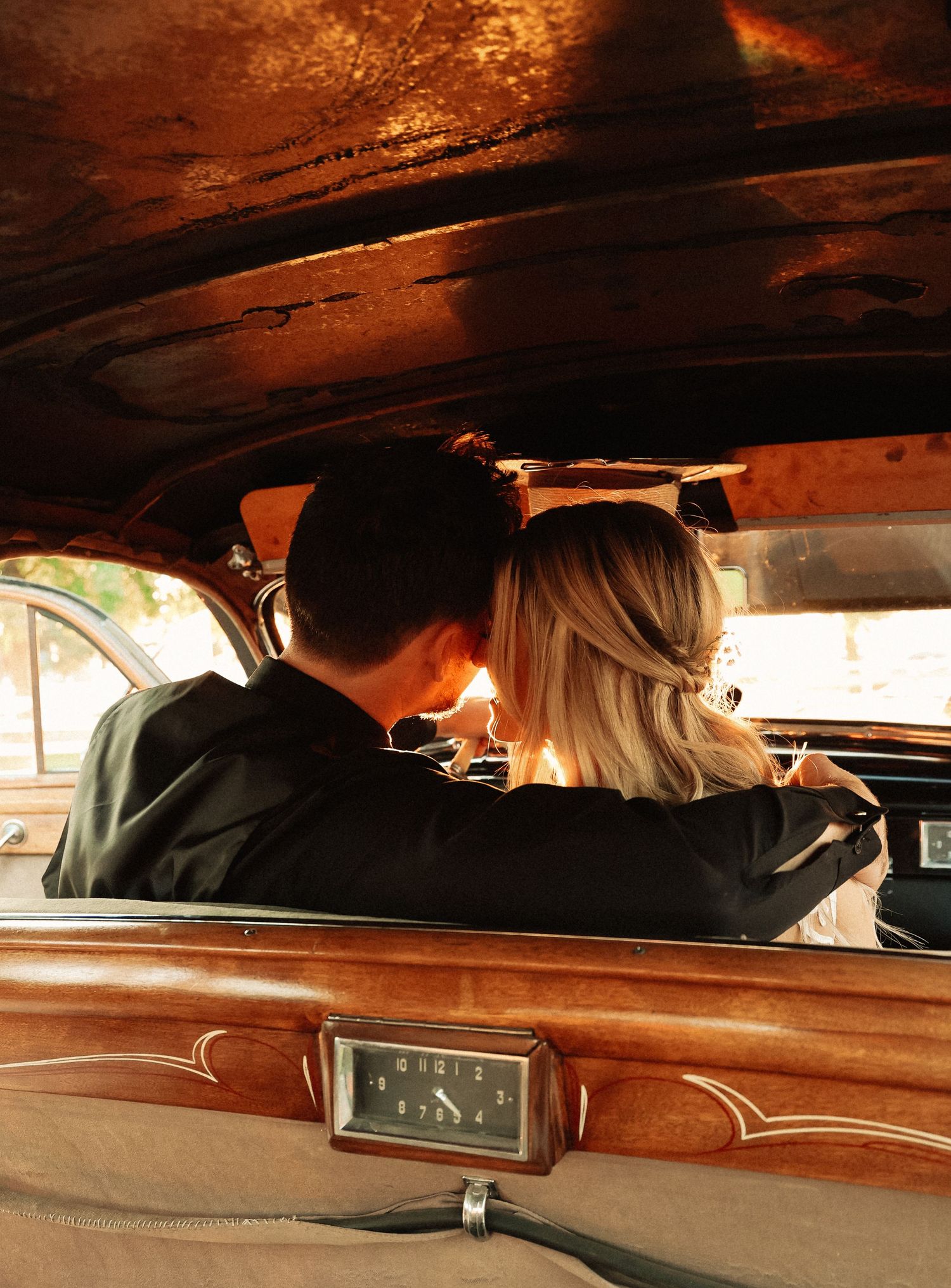 Couple sitting together in vintage car interior with warm amber lighting and wooden dashboard details.