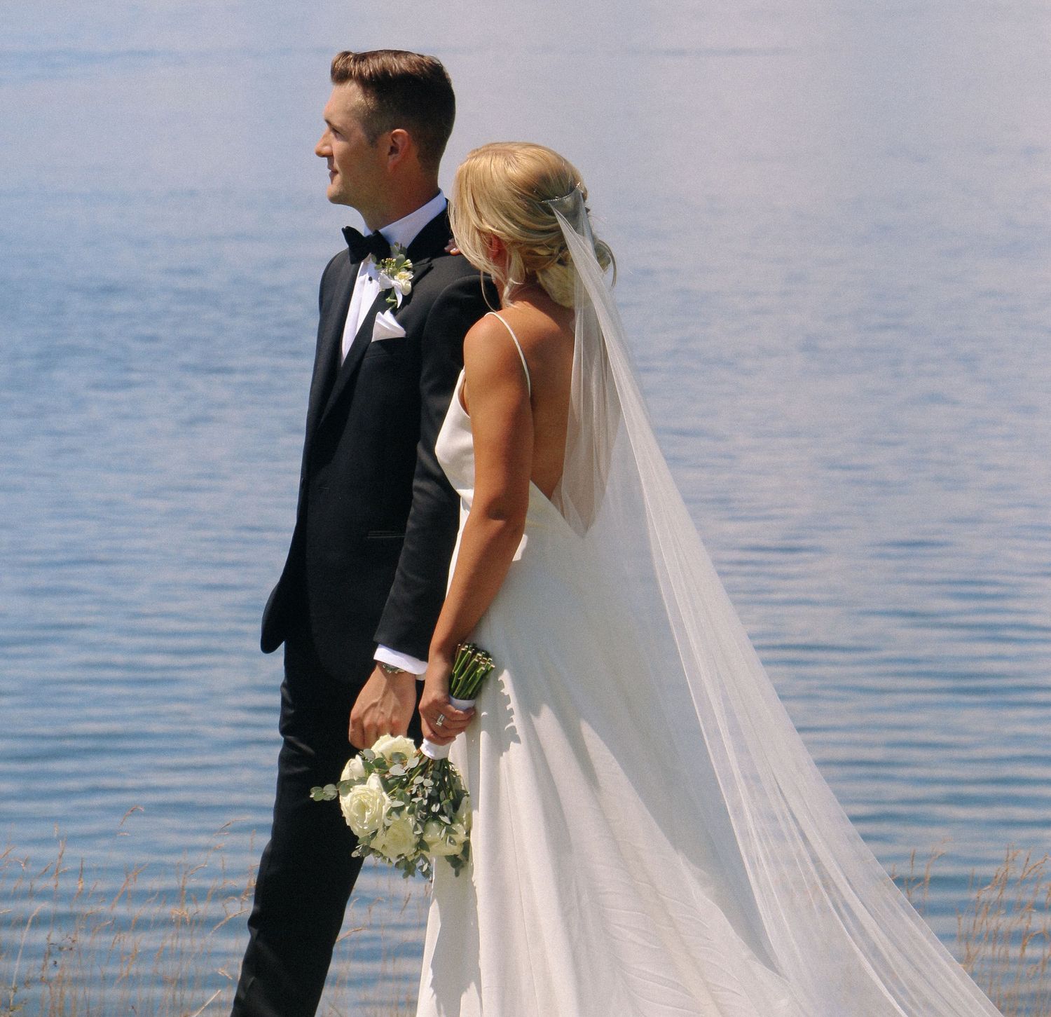 Elegant waterfront wedding photoshoot with flowing white veil against blue water.