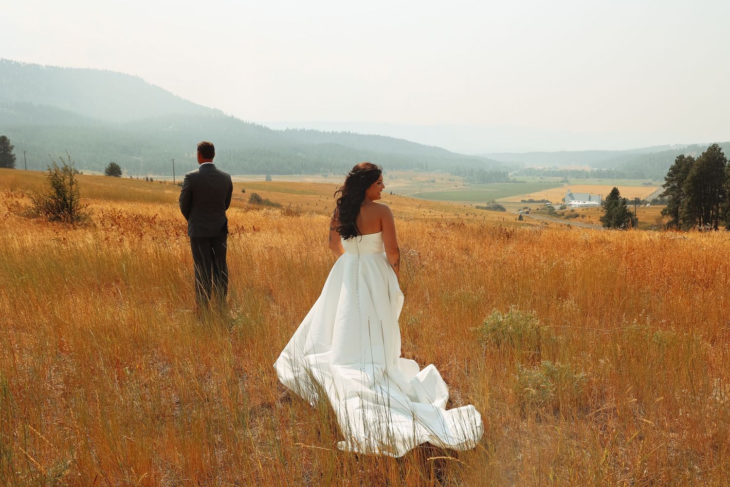 Couple walking through golden field with mountains in background during sunset.