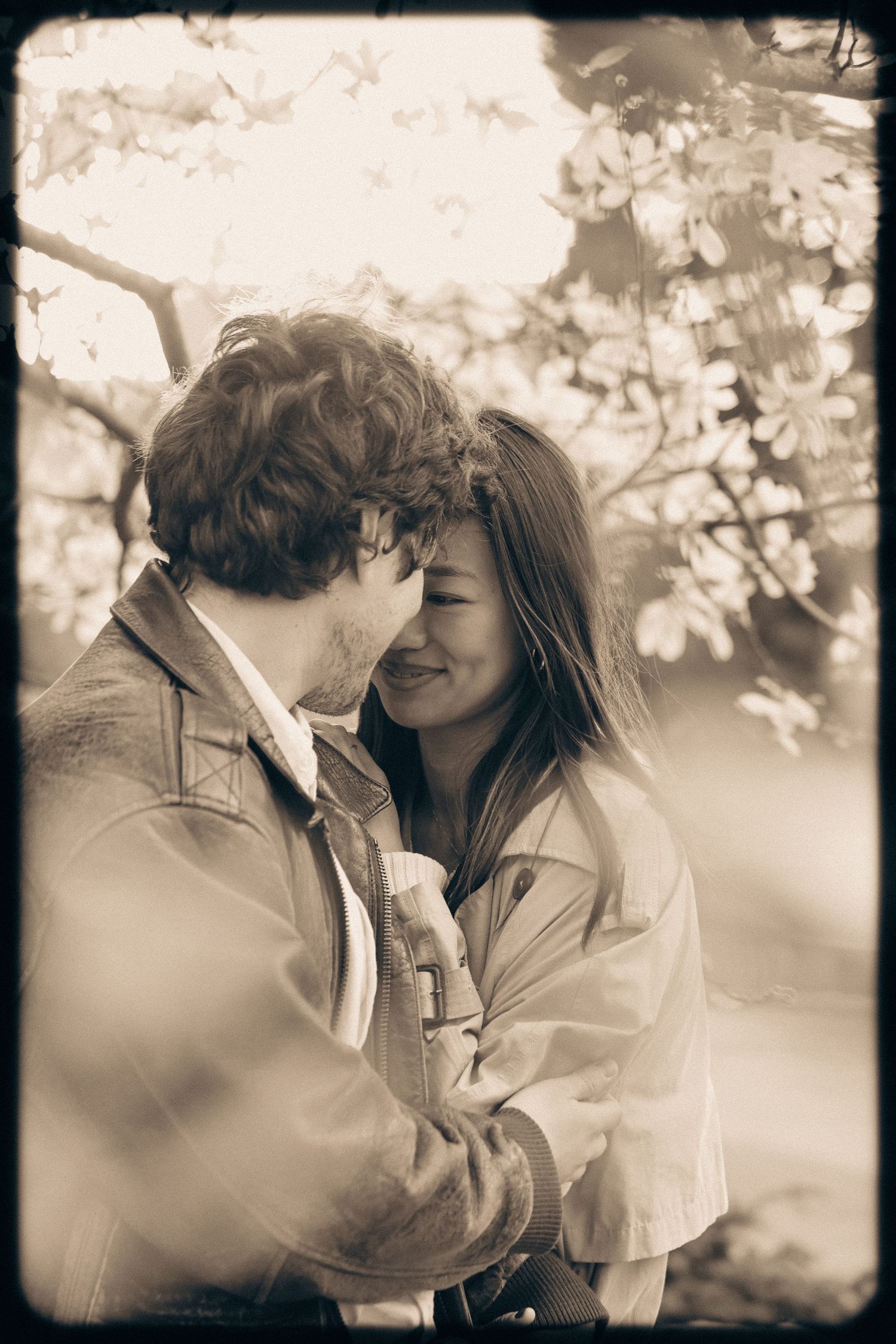 Romantic sepia-toned photo of a couple embracing and smiling under blooming trees in a tender moment.