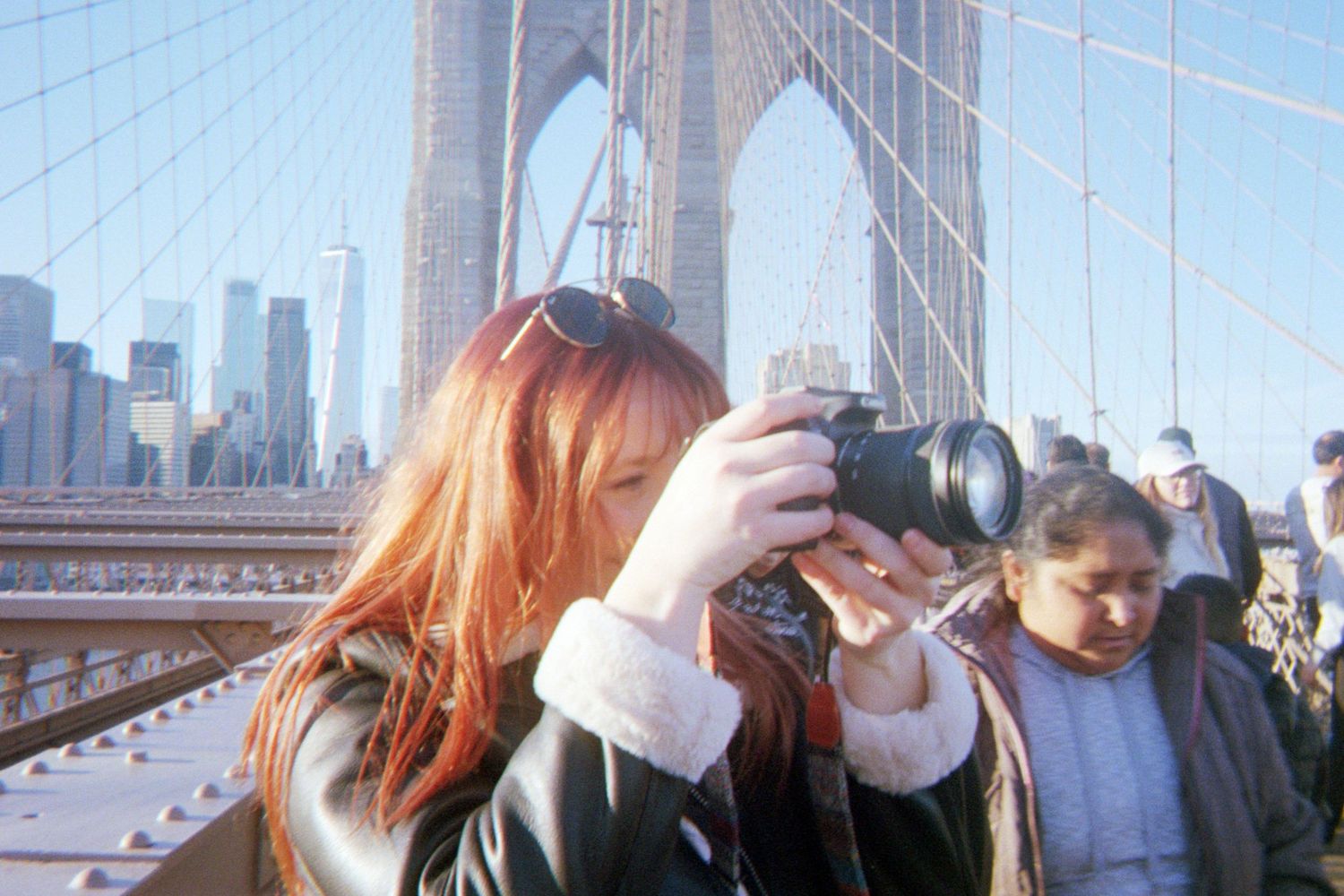 Woman with red hair photographing on Brooklyn Bridge with NYC skyline in background on a sunny day.