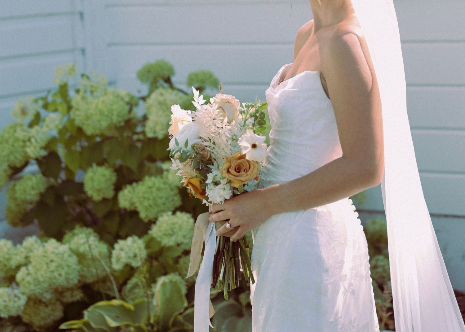 Bride in white strapless wedding gown holding a bouquet of peach and white flowers with ribbon streamers outdoors.