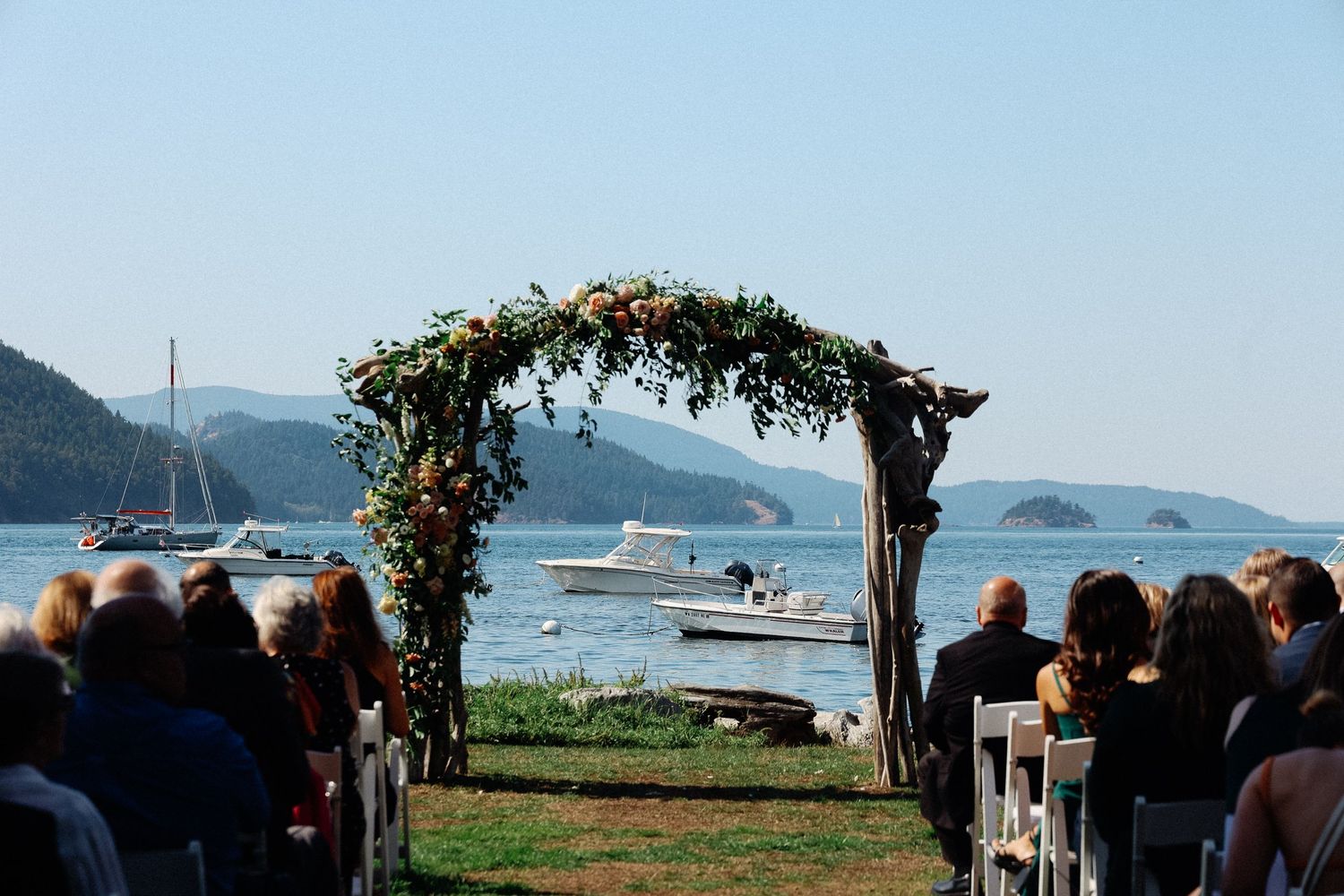 Outdoor waterfront wedding ceremony with floral arch, white chairs, and sailboats on calm blue water with mountain backdrop.
