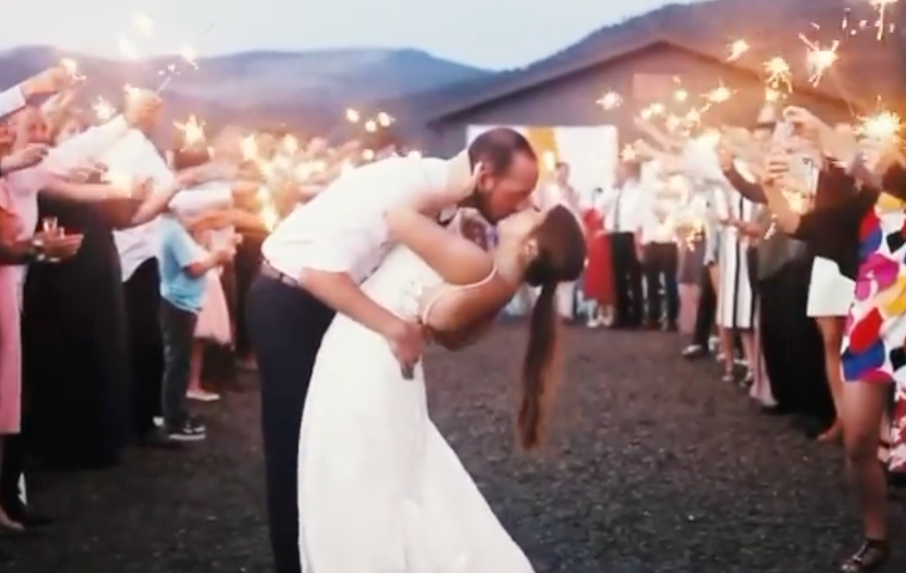 Bride and groom share romantic dip kiss surrounded by sparkler send-off at outdoor evening wedding celebration.