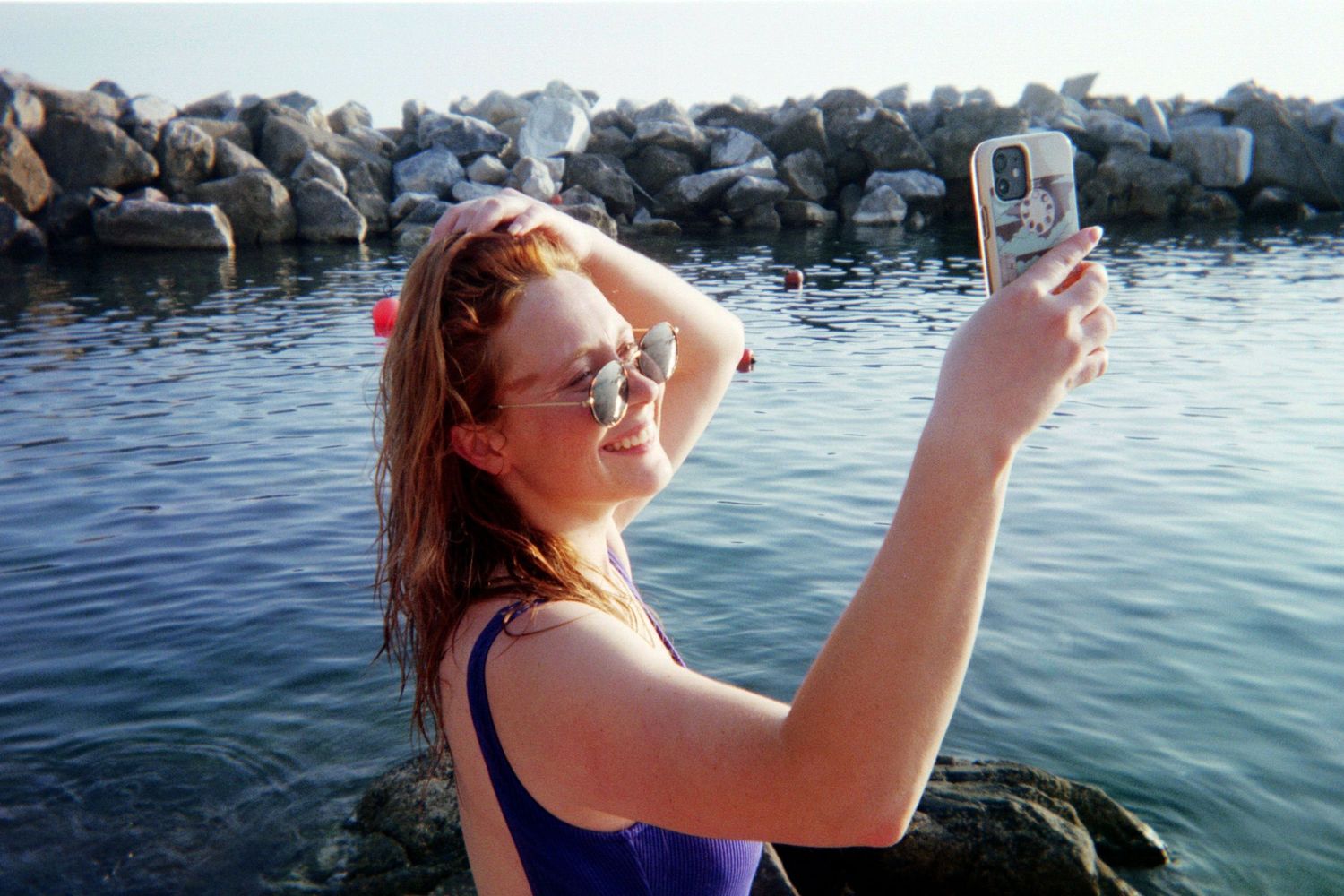 Young woman in purple swimsuit and sunglasses taking a selfie by the rocky seaside on a sunny day.