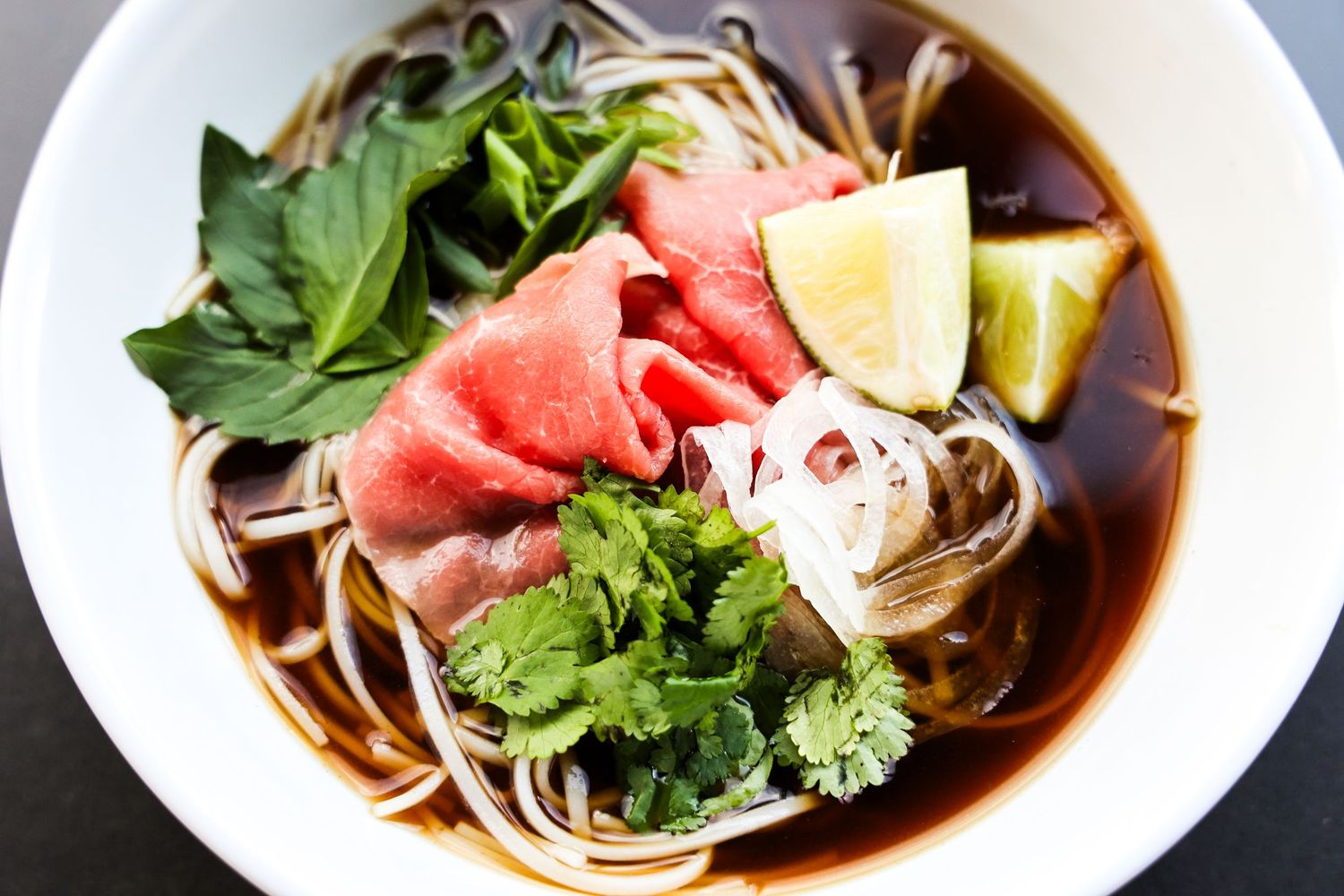 Asian noodle soups garnished with fresh herbs and lime in white bowls.