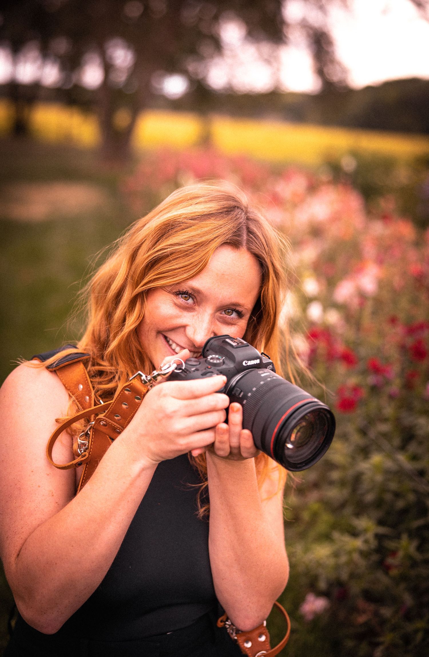 Photographer with wavy red hair smiles while holding a professional camera in a garden with pink flowers at sunset.