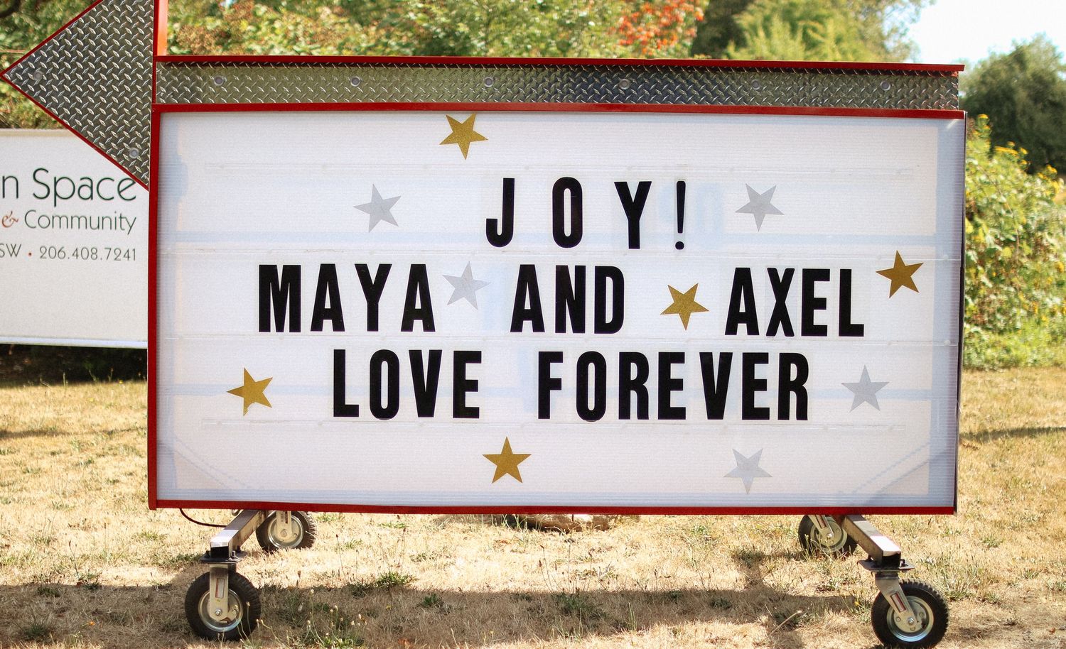 A marquee sign reading JOY! Maya and Axel Love Forever displayed outdoors at a wedding celebration.