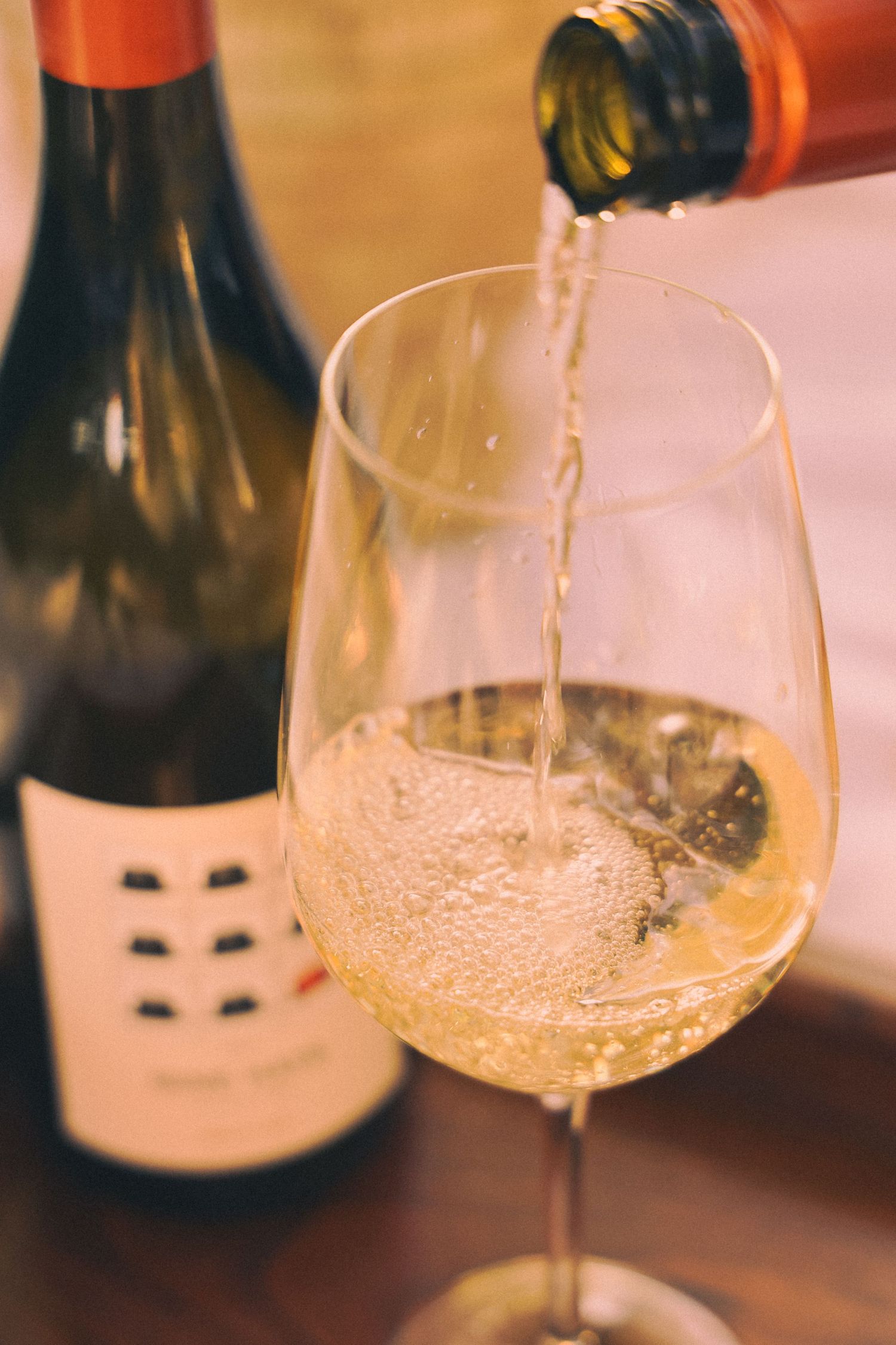 White wine being poured into a crystal glass with wine bottles in the background on a wooden table.