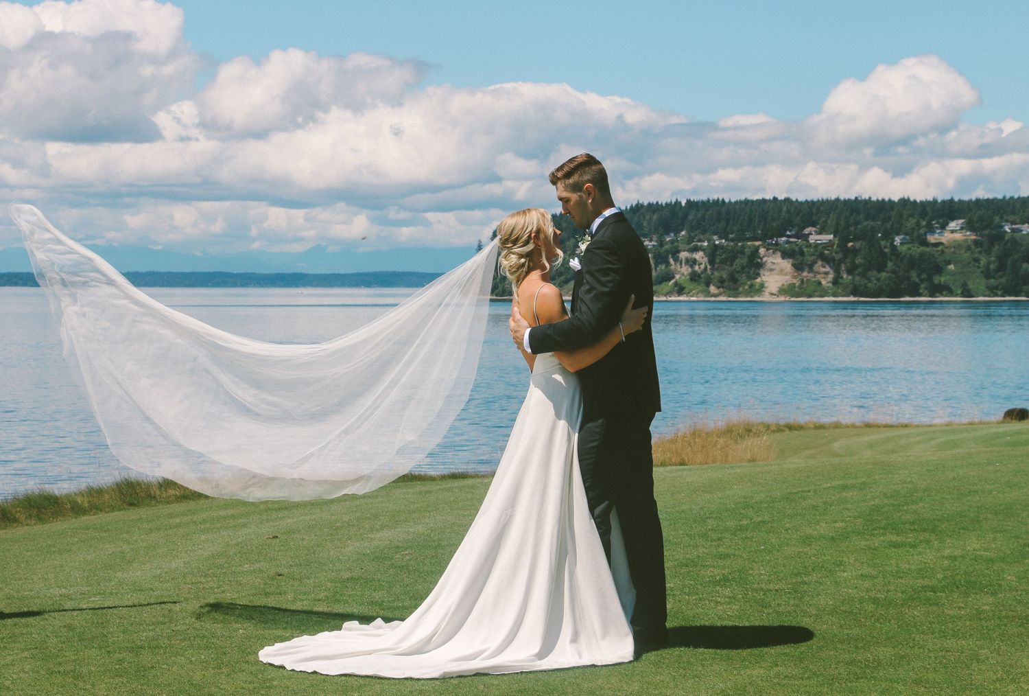 Bride and groom embrace on scenic coastal cliff with flowing veil.