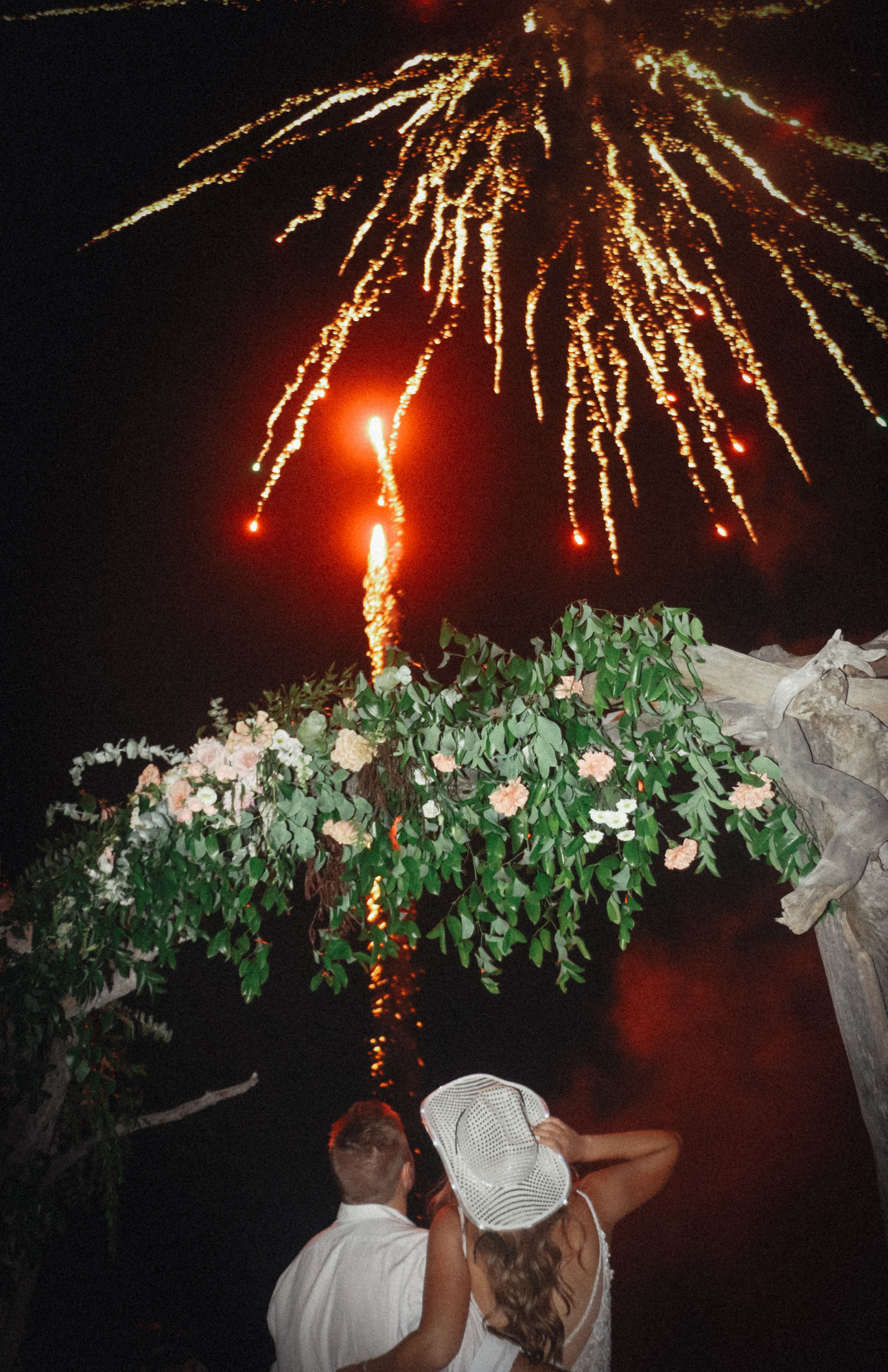 Couple watching fireworks display over floral arch at night celebration.