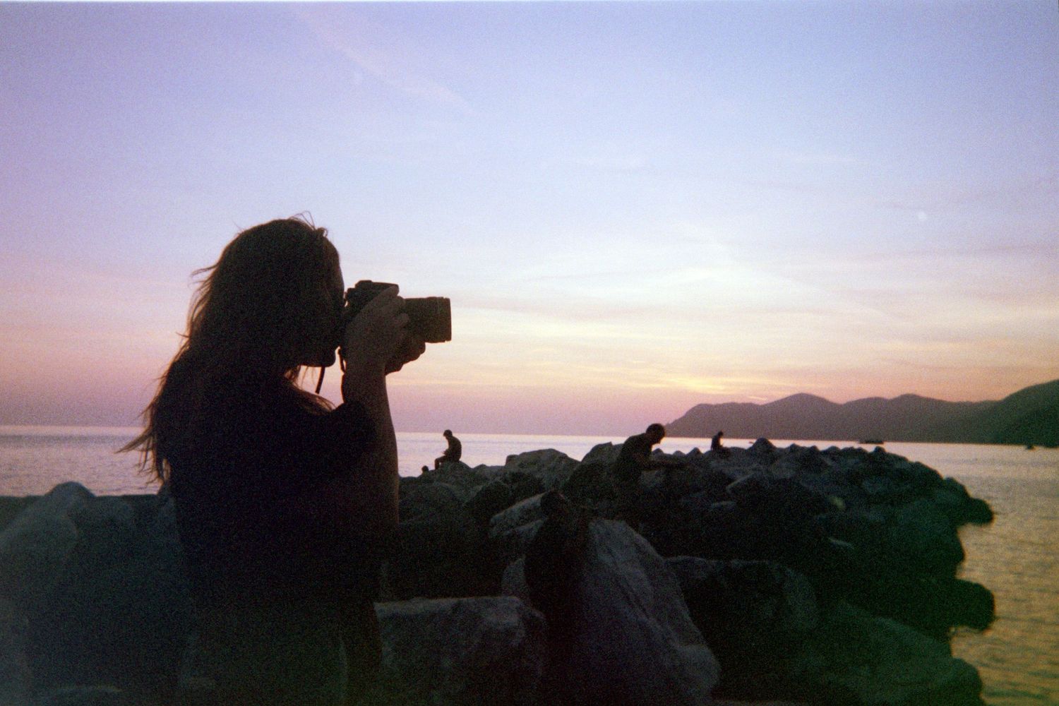 Silhouette of a woman photographing a scenic sunset over rocky coastline with mountains in the background.
