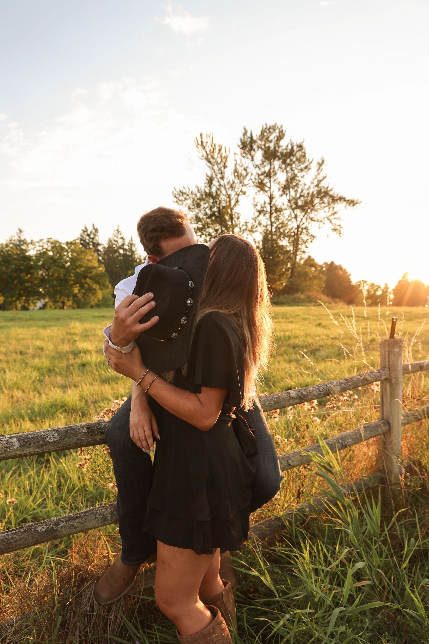 Couple embracing at sunset by a rustic wooden fence in a green meadow, golden hour light glowing behind them.