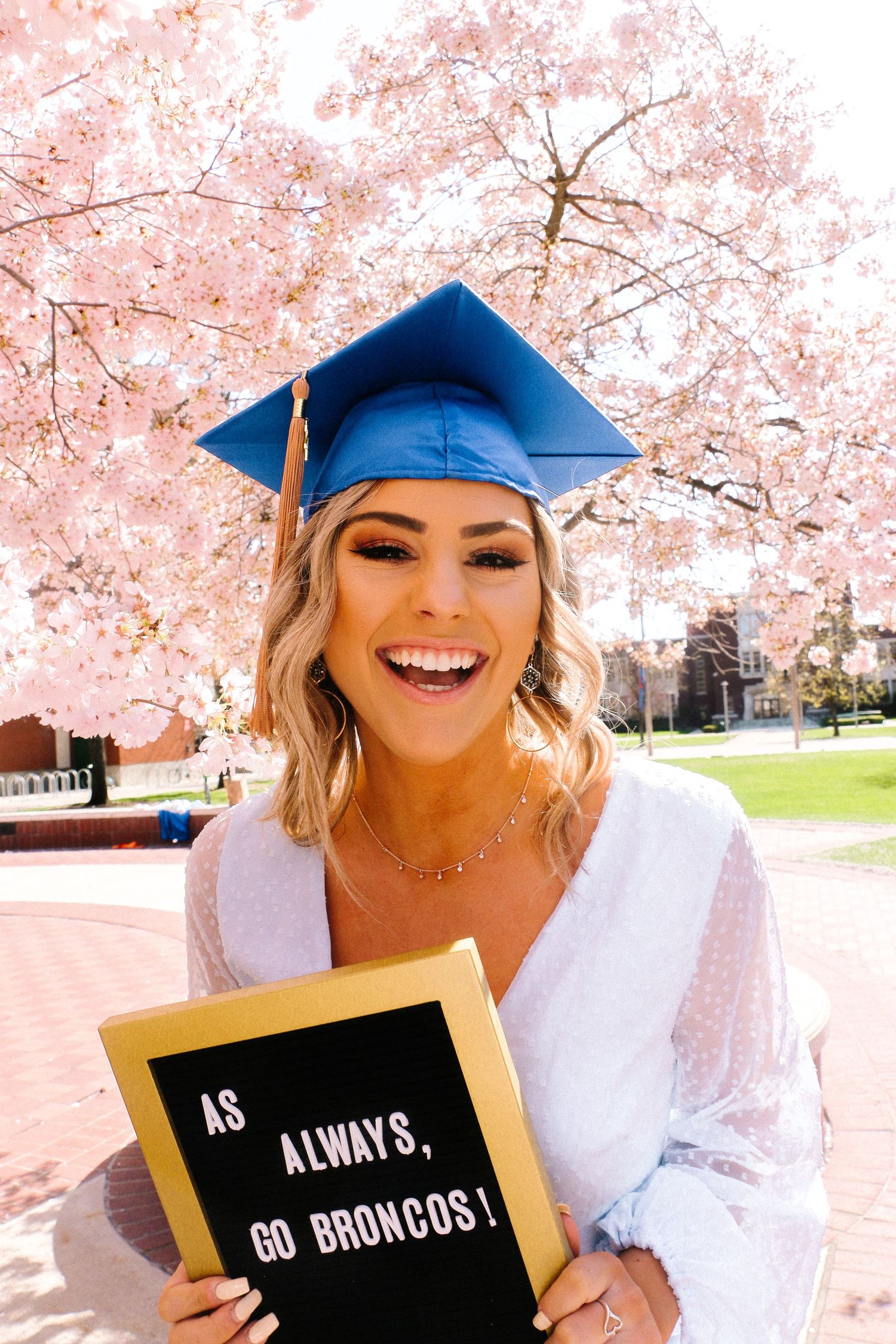 Graduate in blue cap poses with black and gold signs under blooming cherry blossoms on sunny spring day.