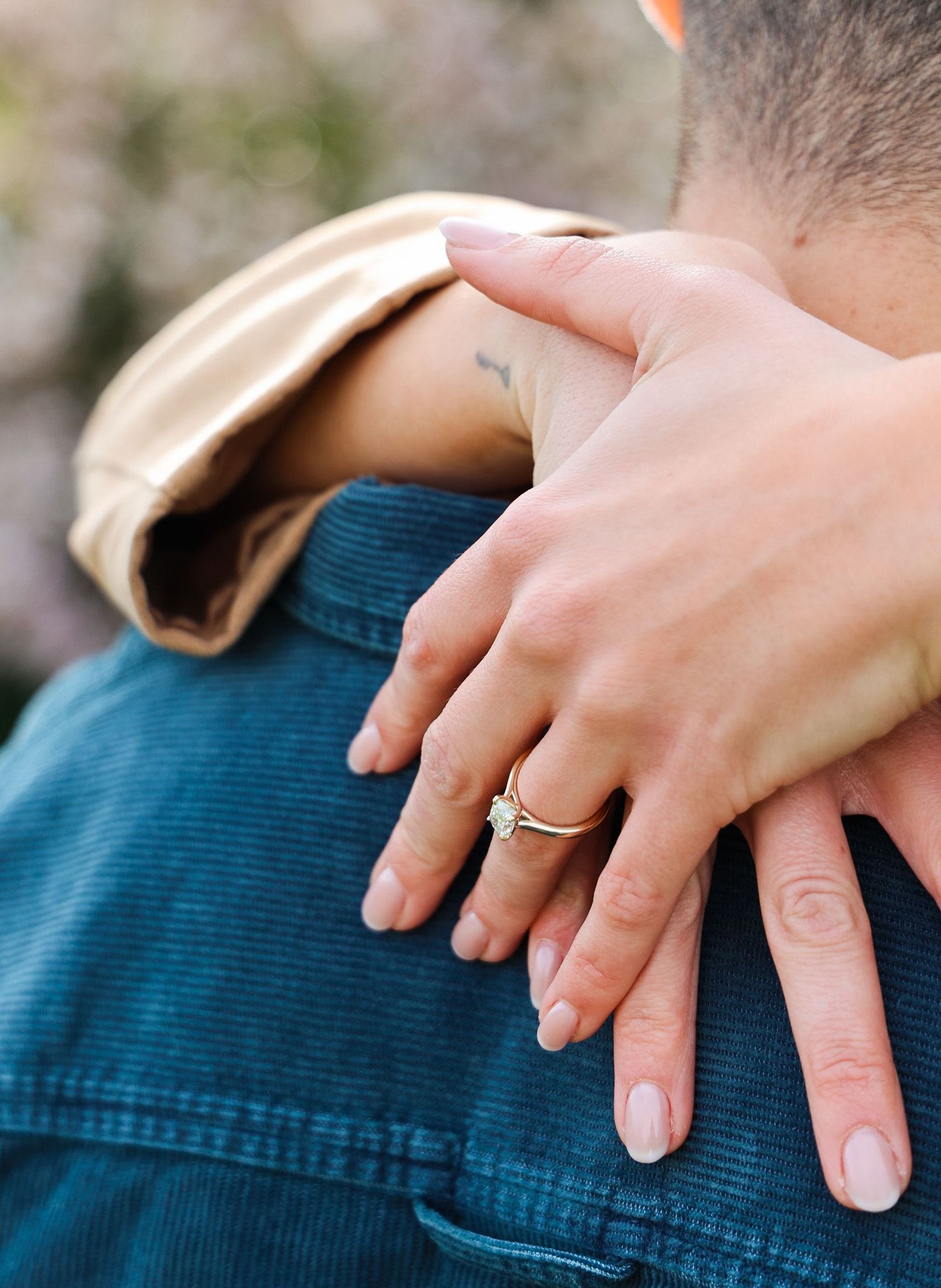 Close-up of a woman's hand with a gold diamond engagement ring resting on a man's shoulder in a teal jacket.