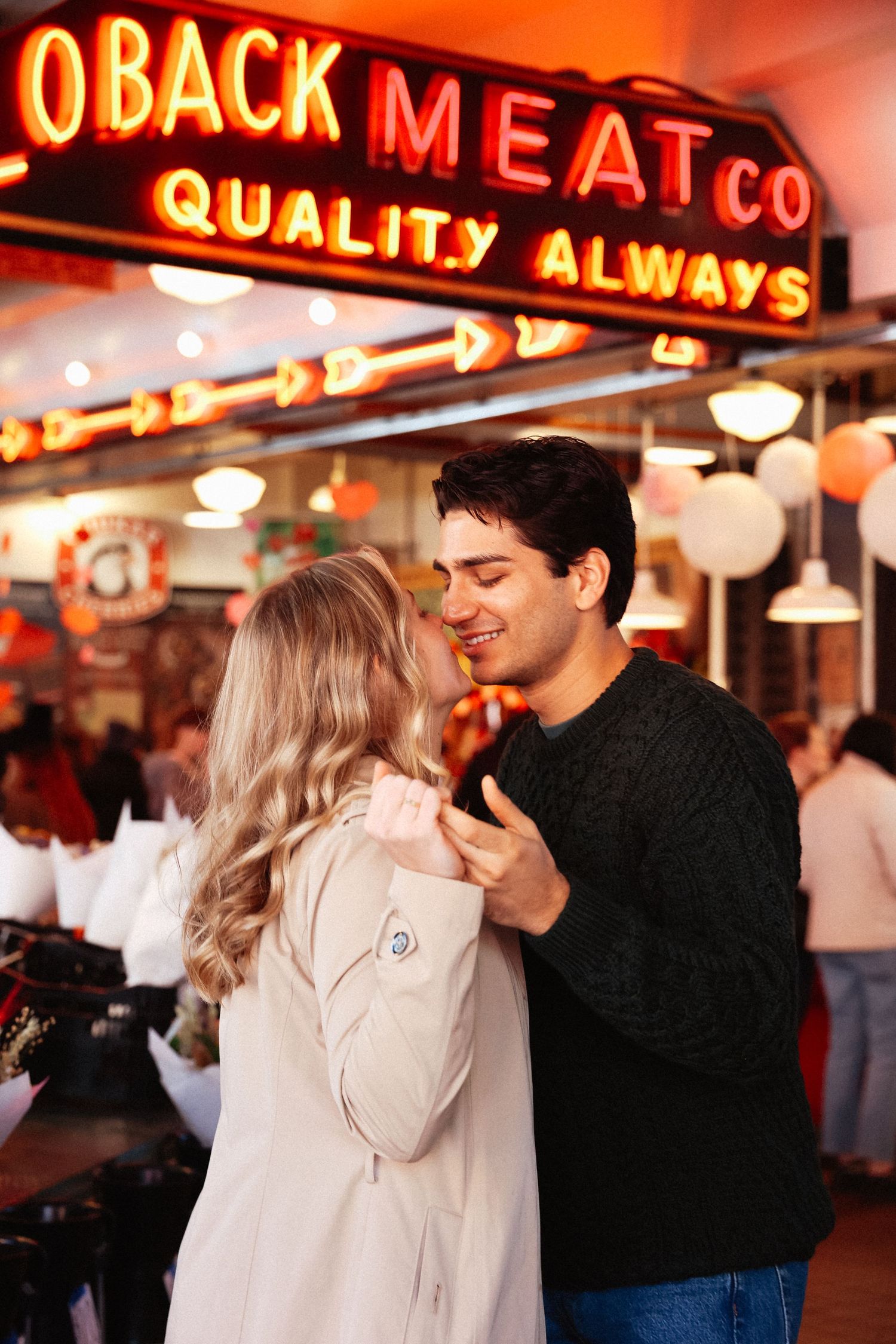 A couple shares an affectionate moment under neon market signs.