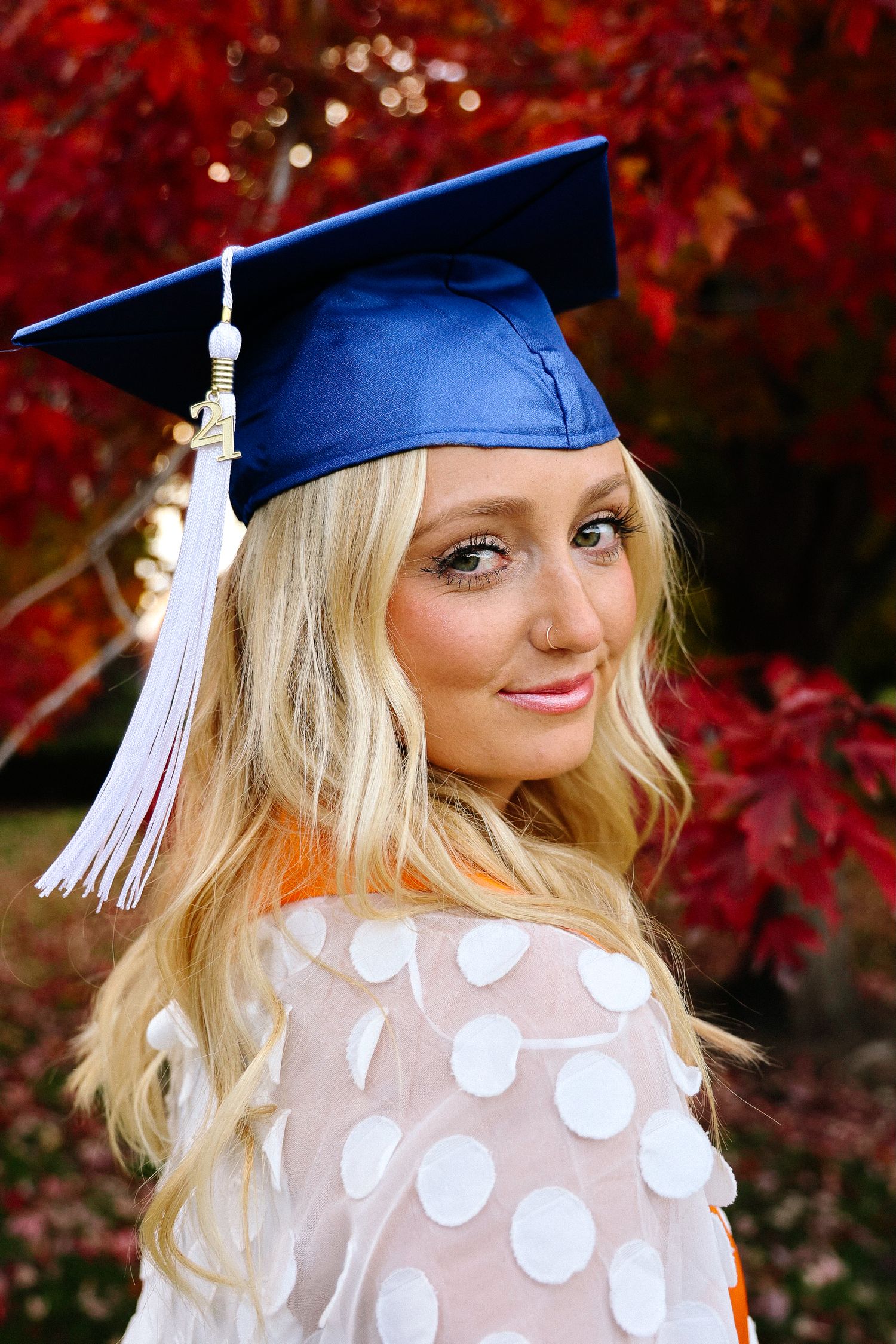 Graduate wearing blue cap and white tassel poses against vibrant red autumn foliage background.