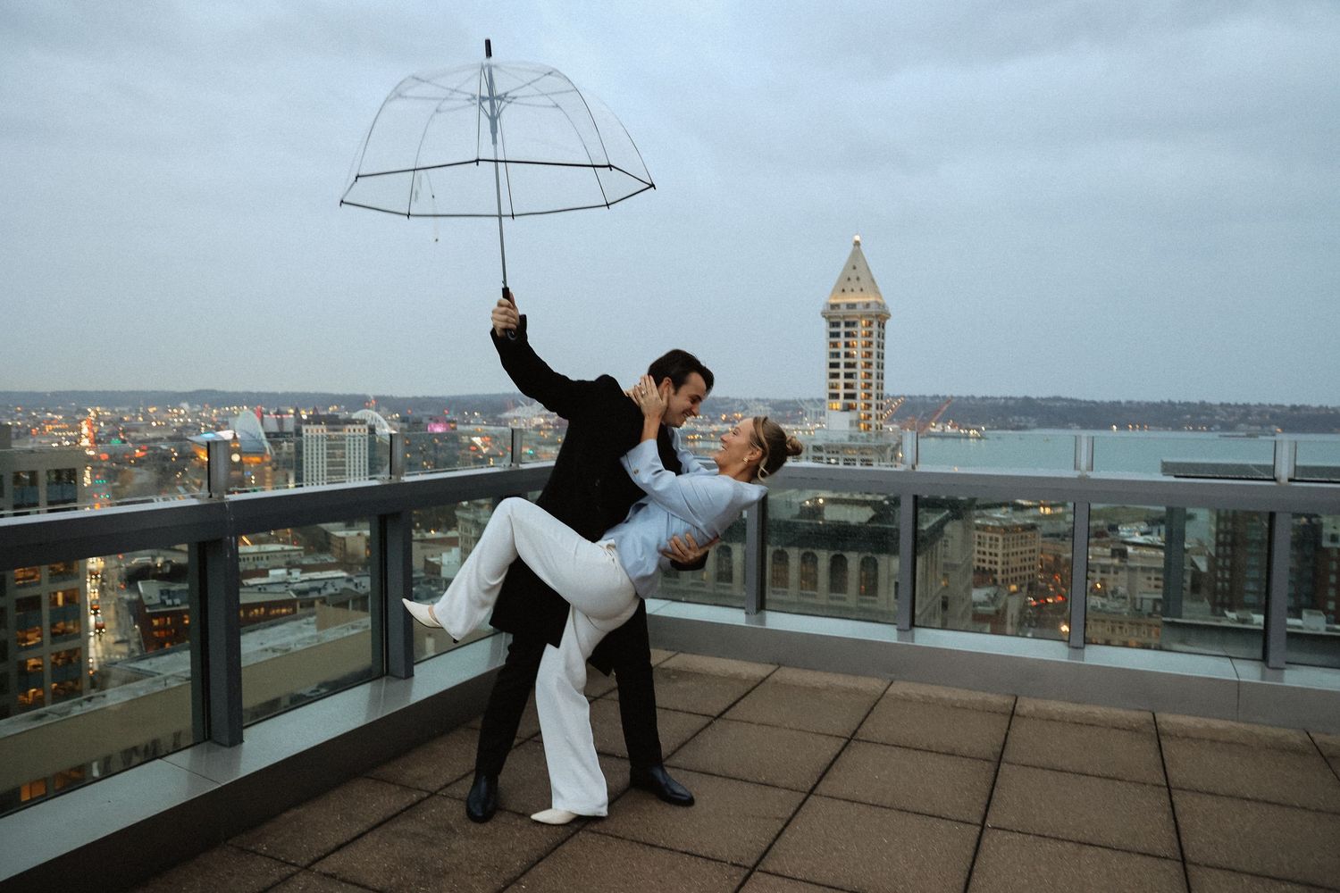 Couple dancing romantically on a rooftop balcony with a clear umbrella against a cityscape.