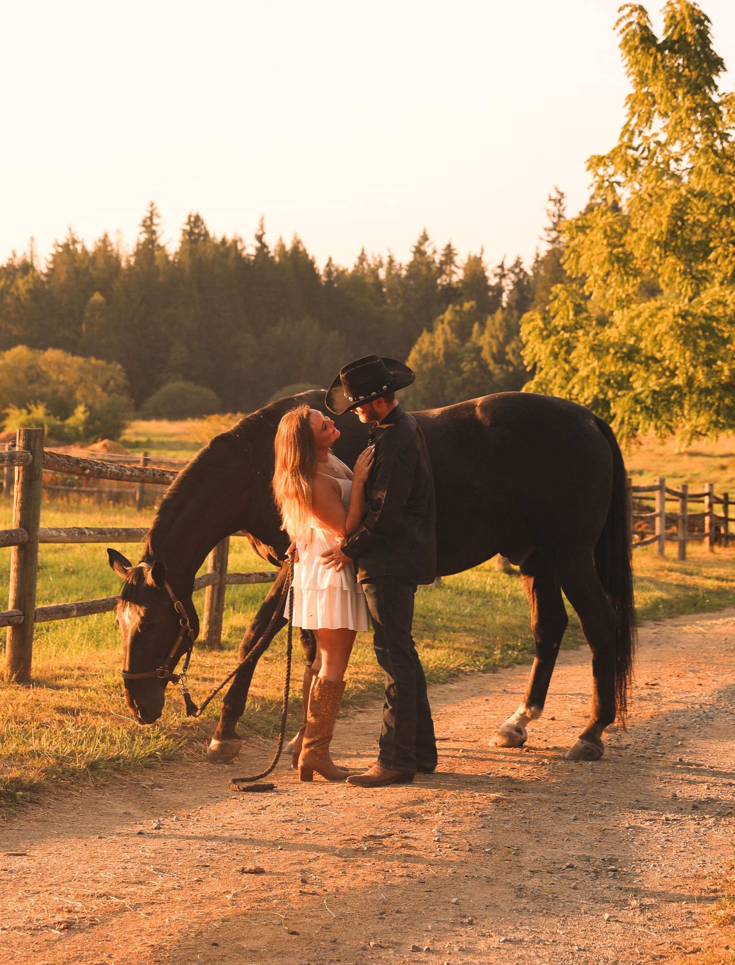 Couple checking horse hooves at sunset on a dirt path with trees in background.