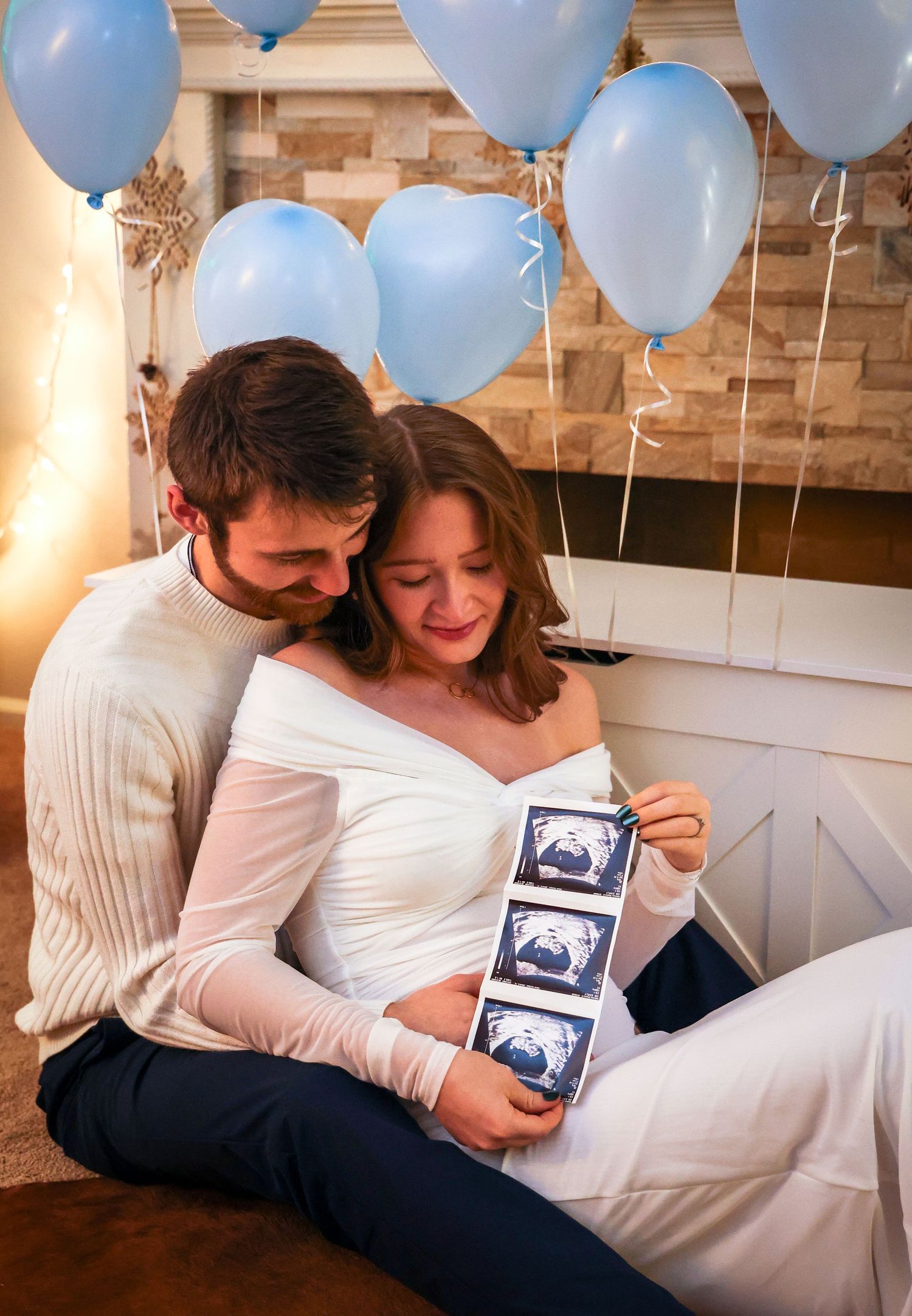 Expecting couple shares tender moment while looking at ultrasound photos surrounded by blue balloons.
