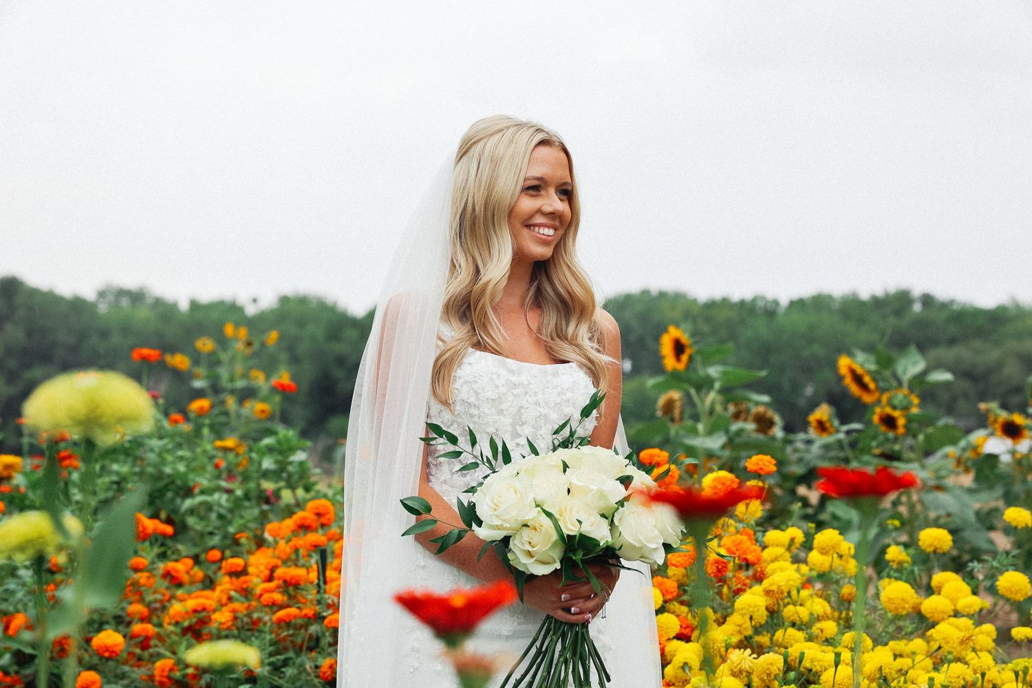 Woman in white dress poses joyfully in vibrant field of orange and yellow flowers.
