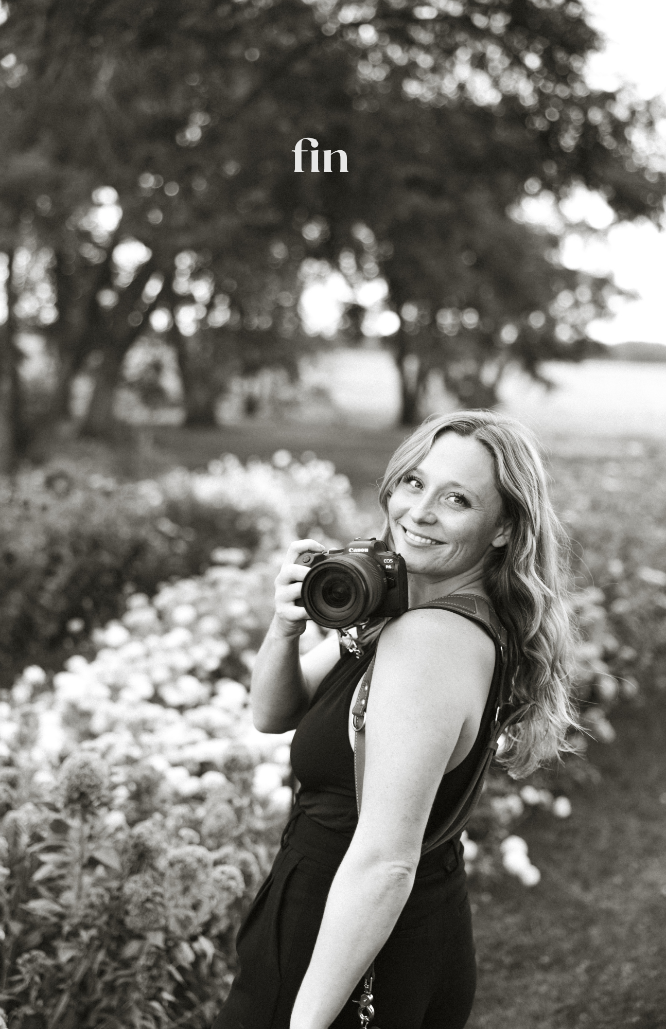 Smiling woman holding a DSLR camera outdoors in a garden, black and white portrait photo with text fin.