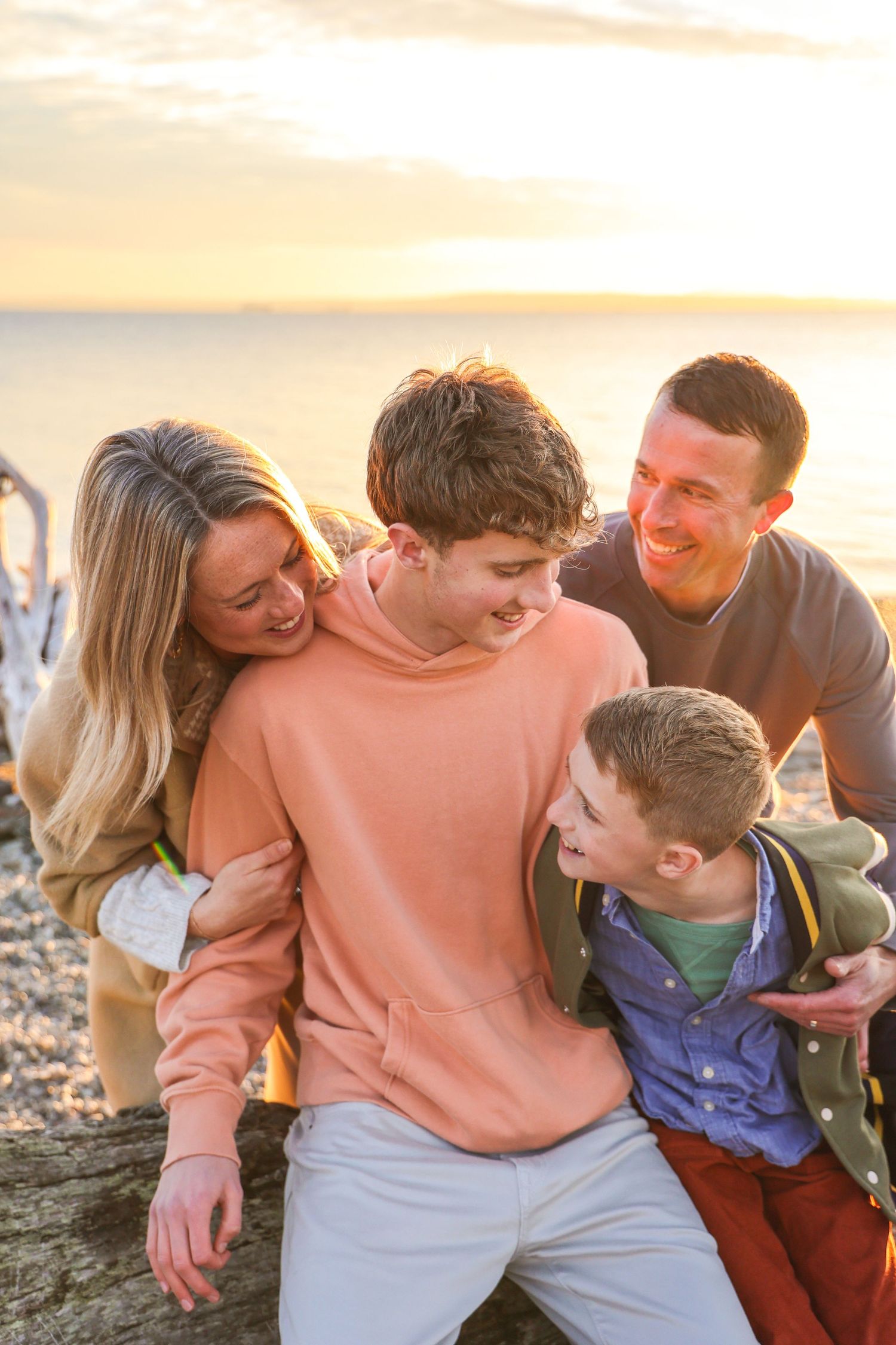 Family of four laughing together on a beach at golden hour sunset, parents and two boys enjoying a warm embrace.