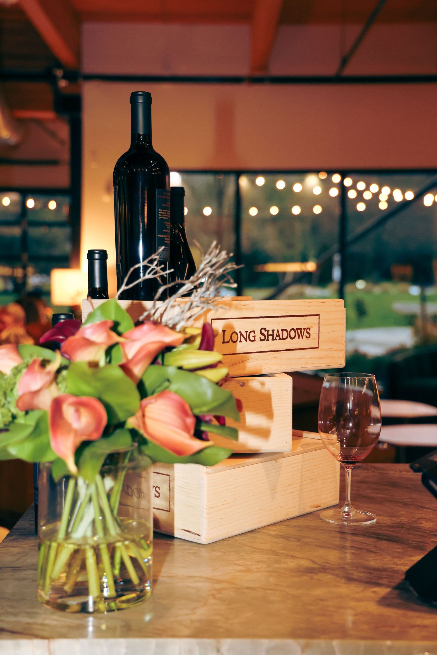 Wine bottles and floral arrangements in pink and green sit on a rustic wooden table at a romantic restaurant setting.