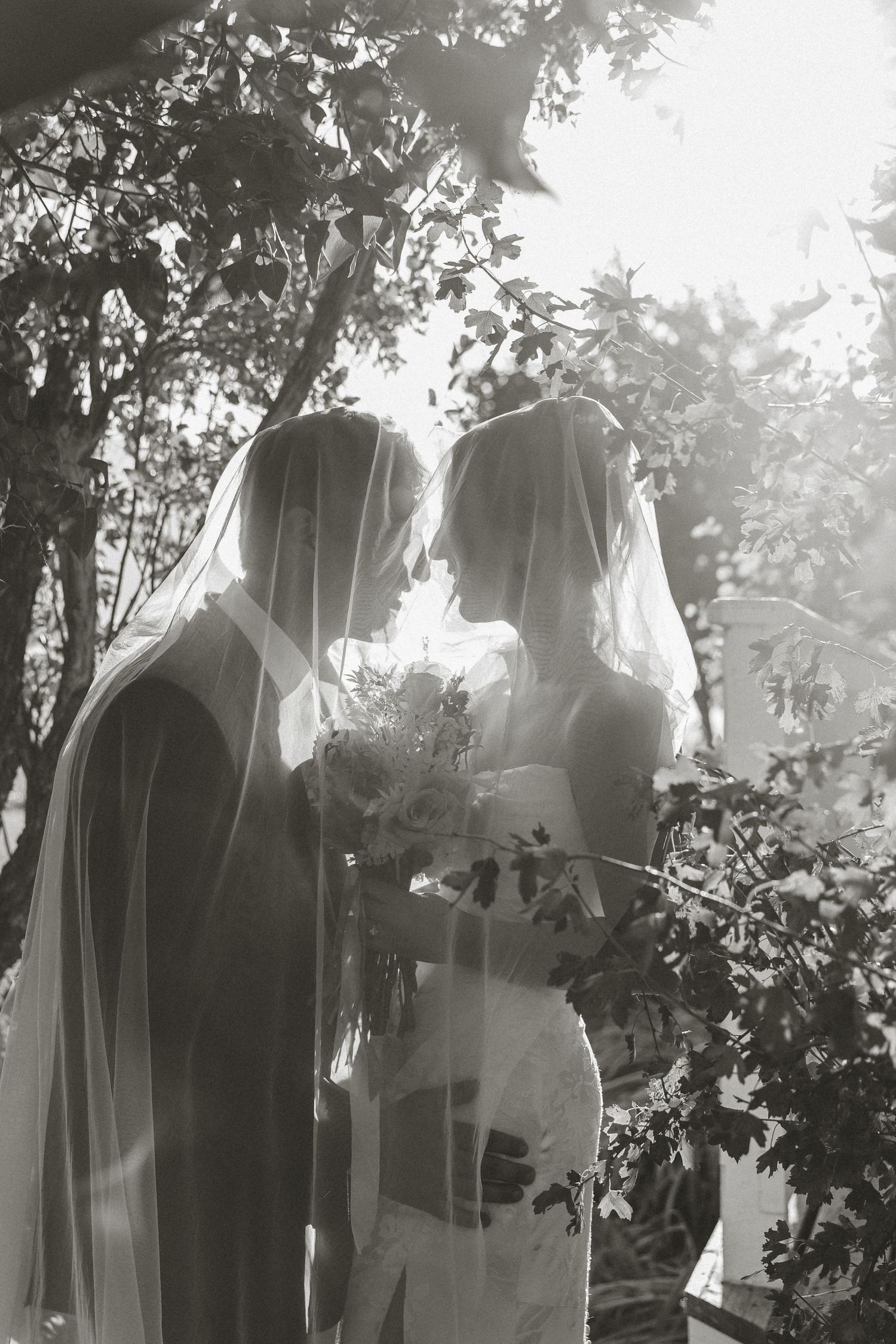 A black and white wedding photo of a couple beneath their veils in a garden setting.