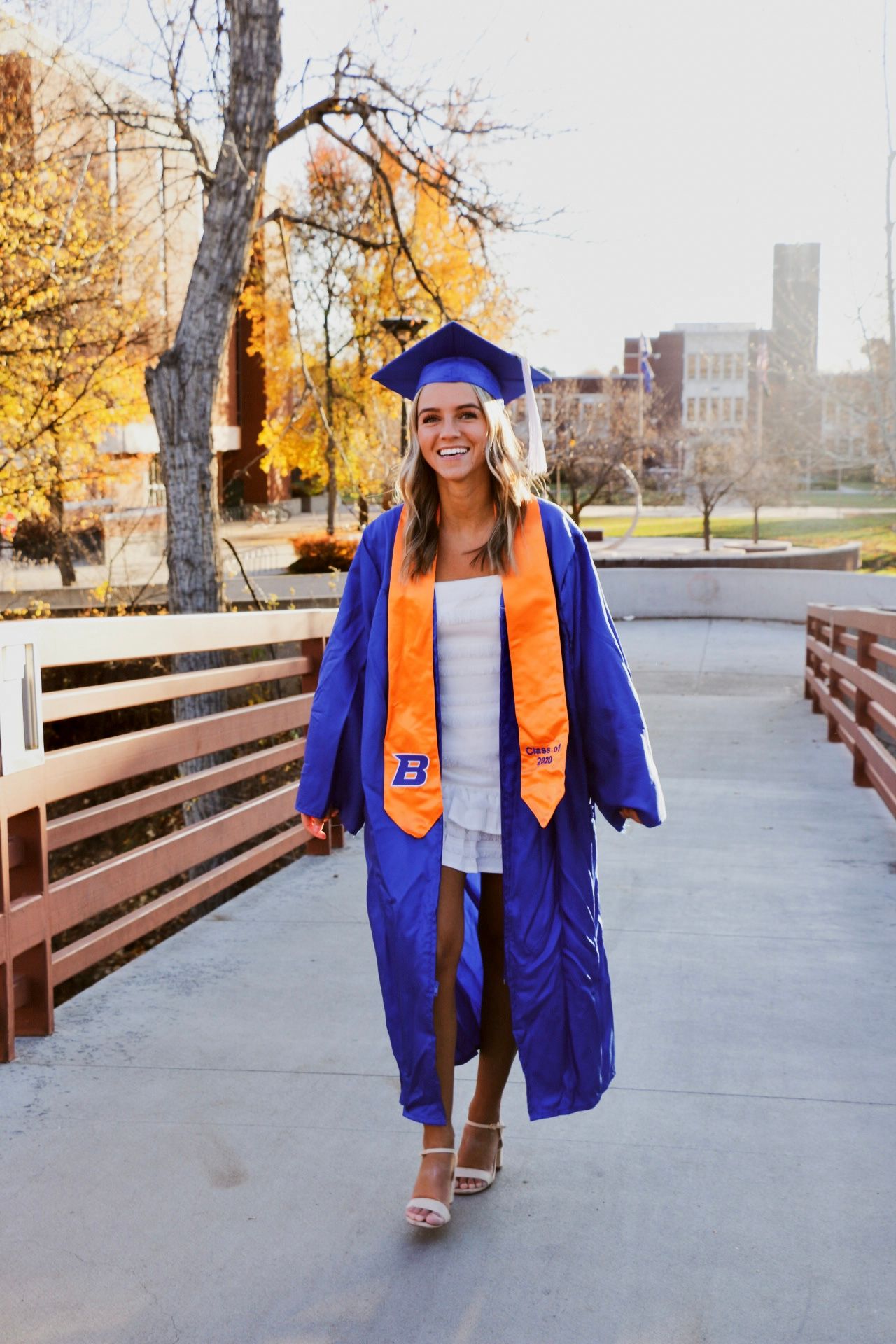 Graduate in blue cap and gown with orange stole walking on campus pathway in autumn.