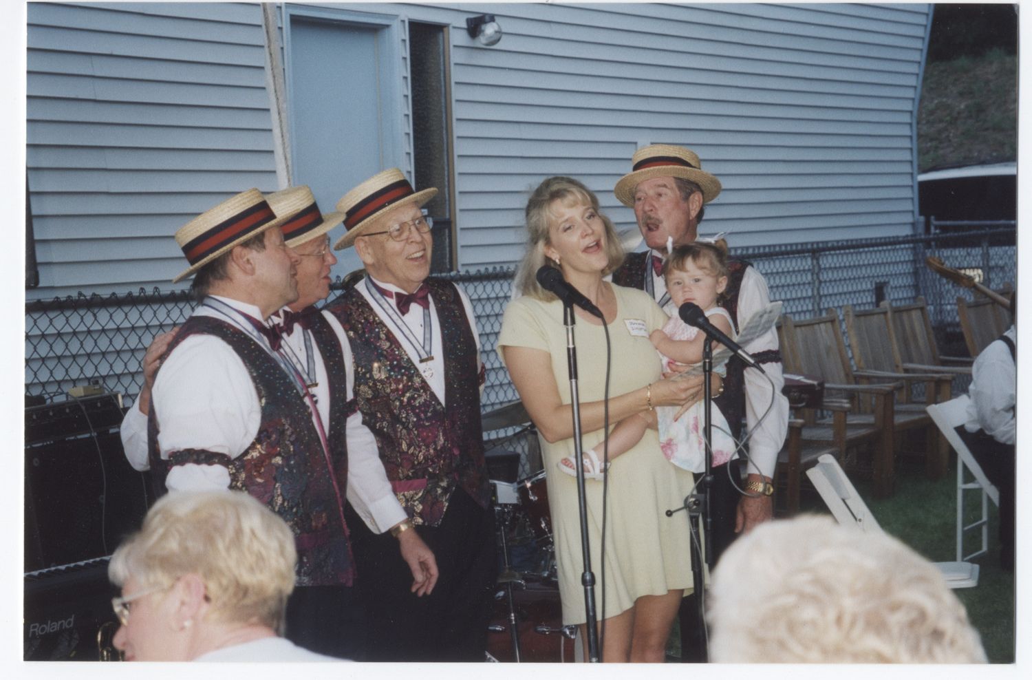 Barbershop quartet in straw hats and vests performing outdoors with a woman singer at a microphone.