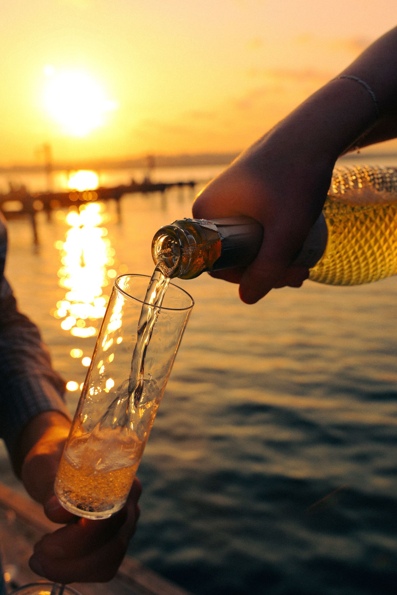 Pouring golden beer into glass at sunset over water with sparkling reflections creating a serene waterfront scene.