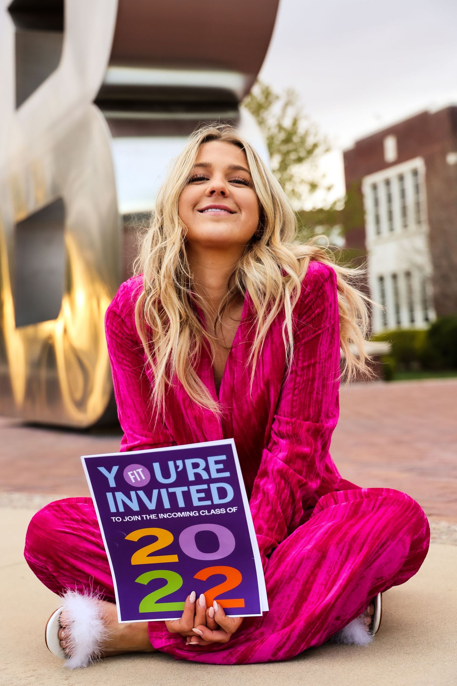 Graduate in bright pink dress lying on brick ground holding acceptance letter.