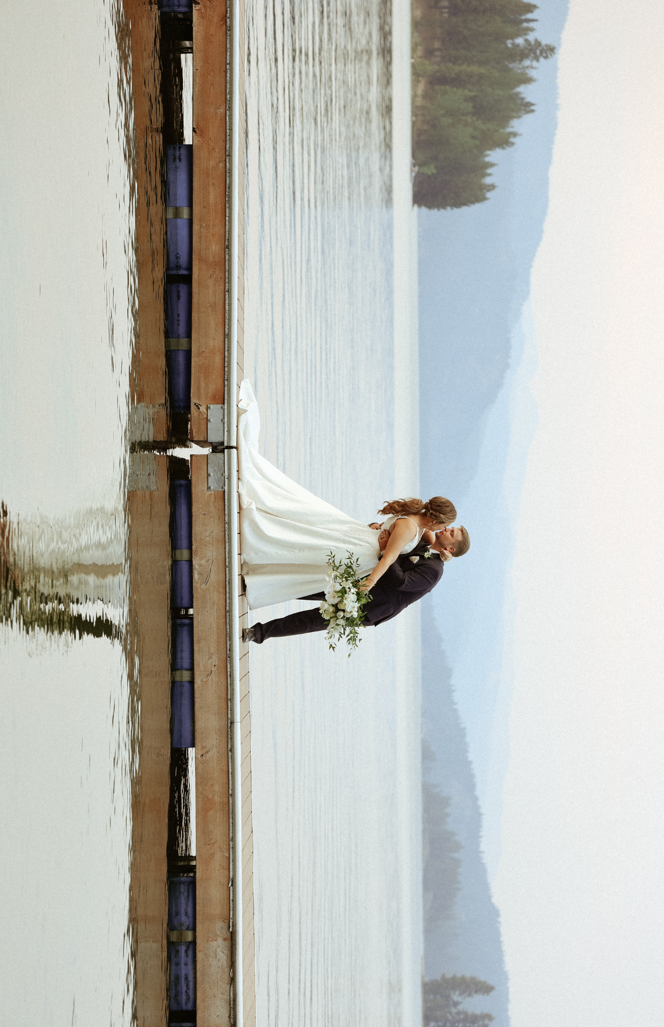 Bride and groom sharing a romantic kiss on a wooden dock beside a serene lake with mountain views on their wedding day.