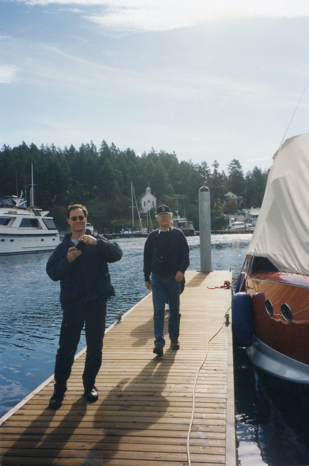 Two men walking on a wooden dock at a marina with boats and tree-lined waterfront in the background.