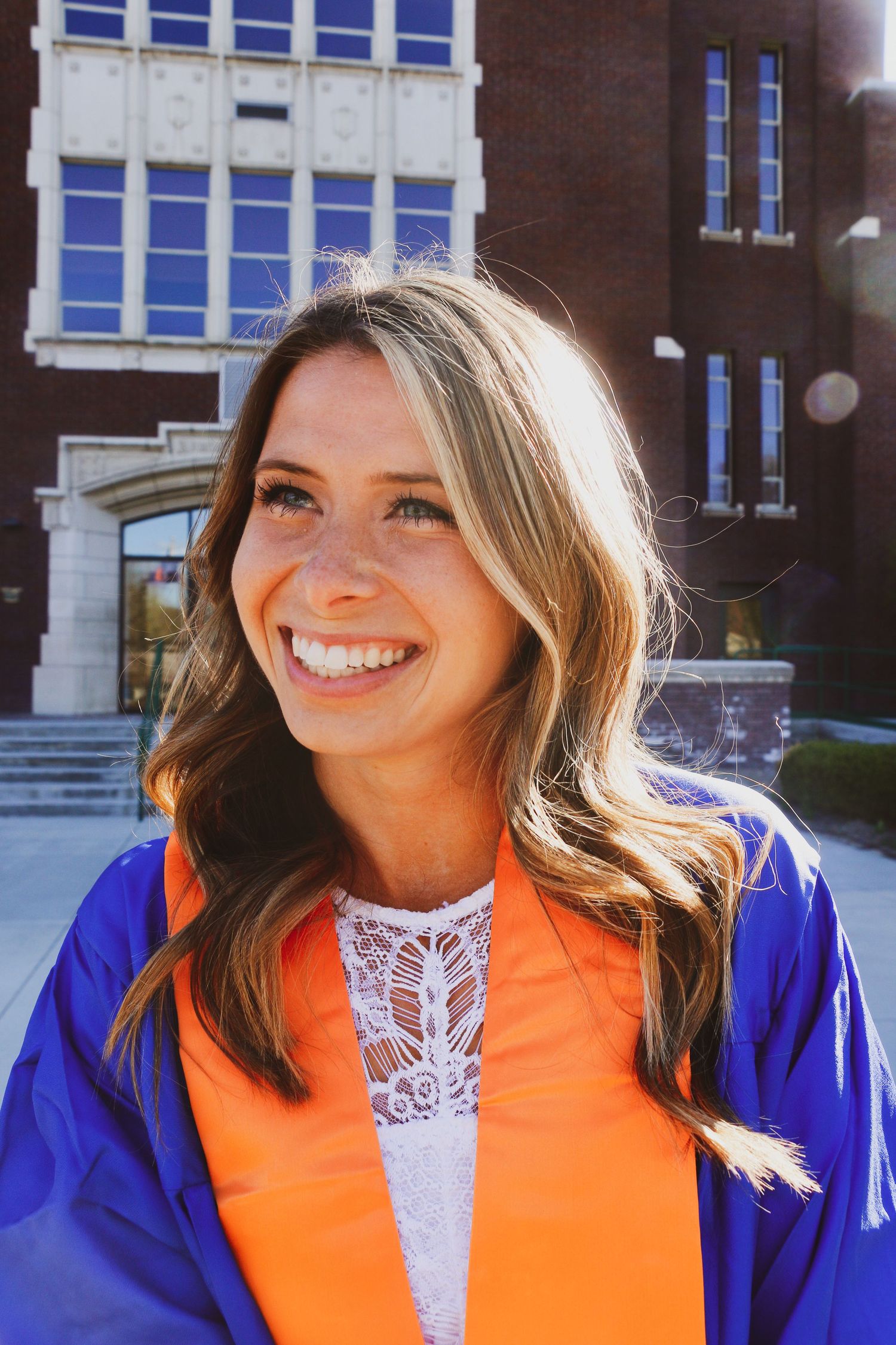 Graduate wearing blue and orange academic regalia smiles brightly on sunny graduation day.