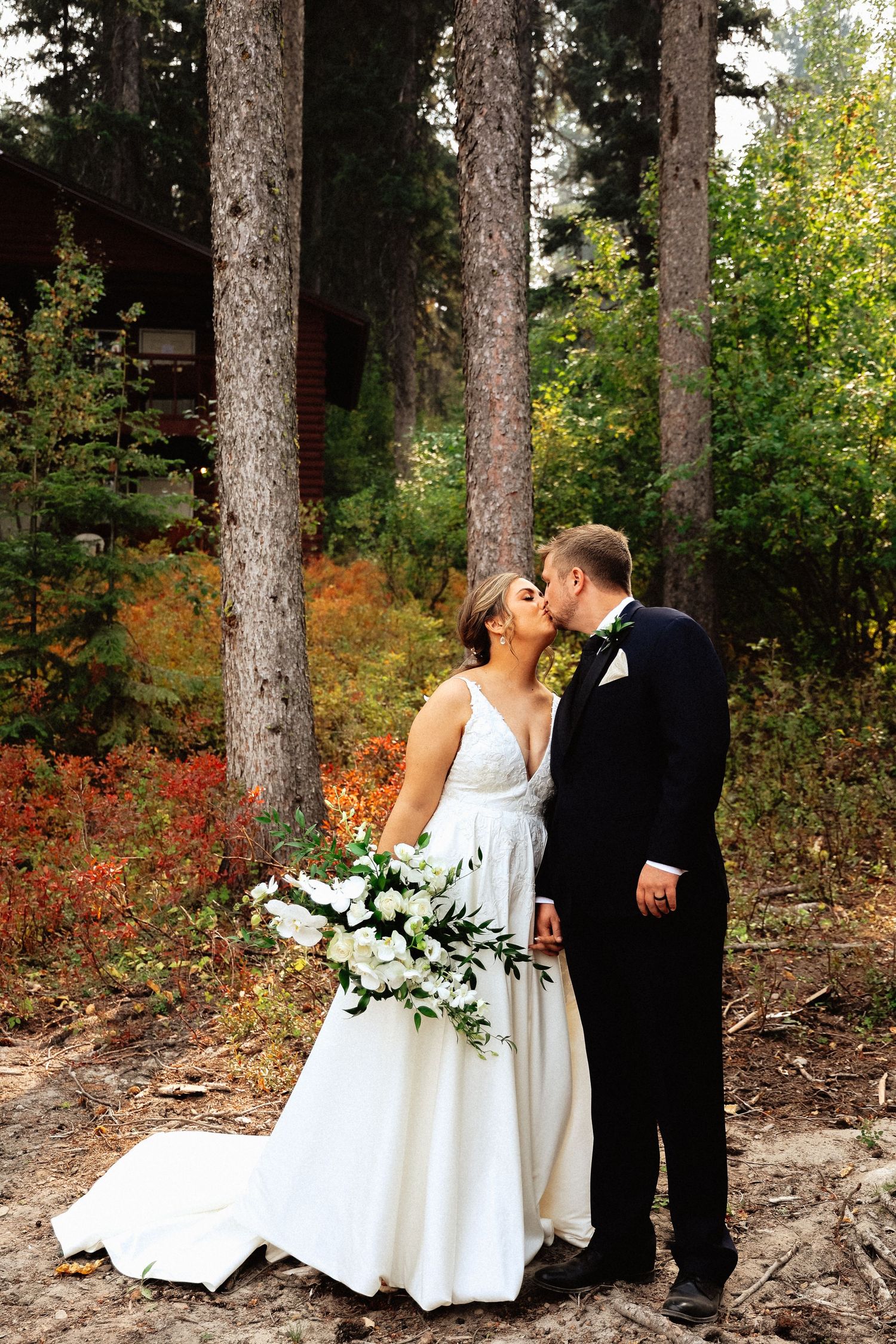 A couple in wedding attire shares a romantic moment among tall pine trees and vibrant autumn foliage.