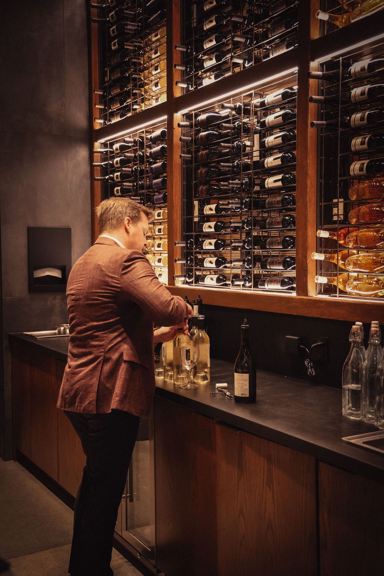 Wine steward organizing bottles in illuminated wall-mounted wine storage display.
