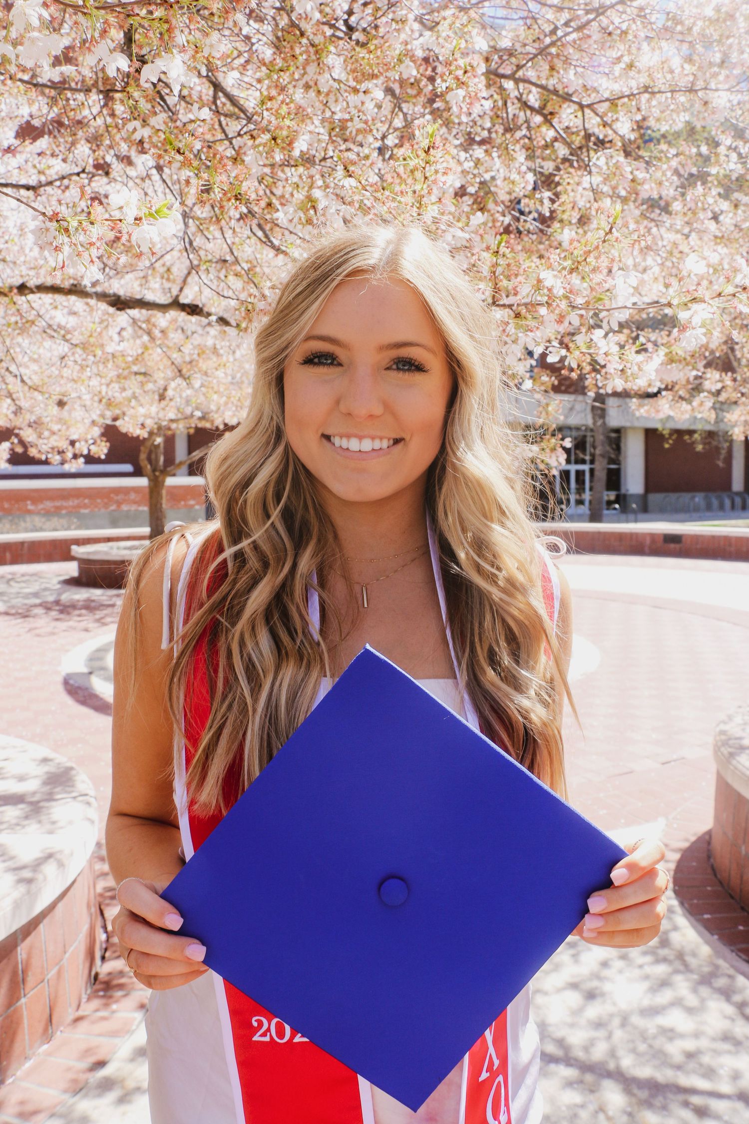 A graduate in a red dress holds a blue graduation cap under blooming cherry blossom trees on a sunny spring day.