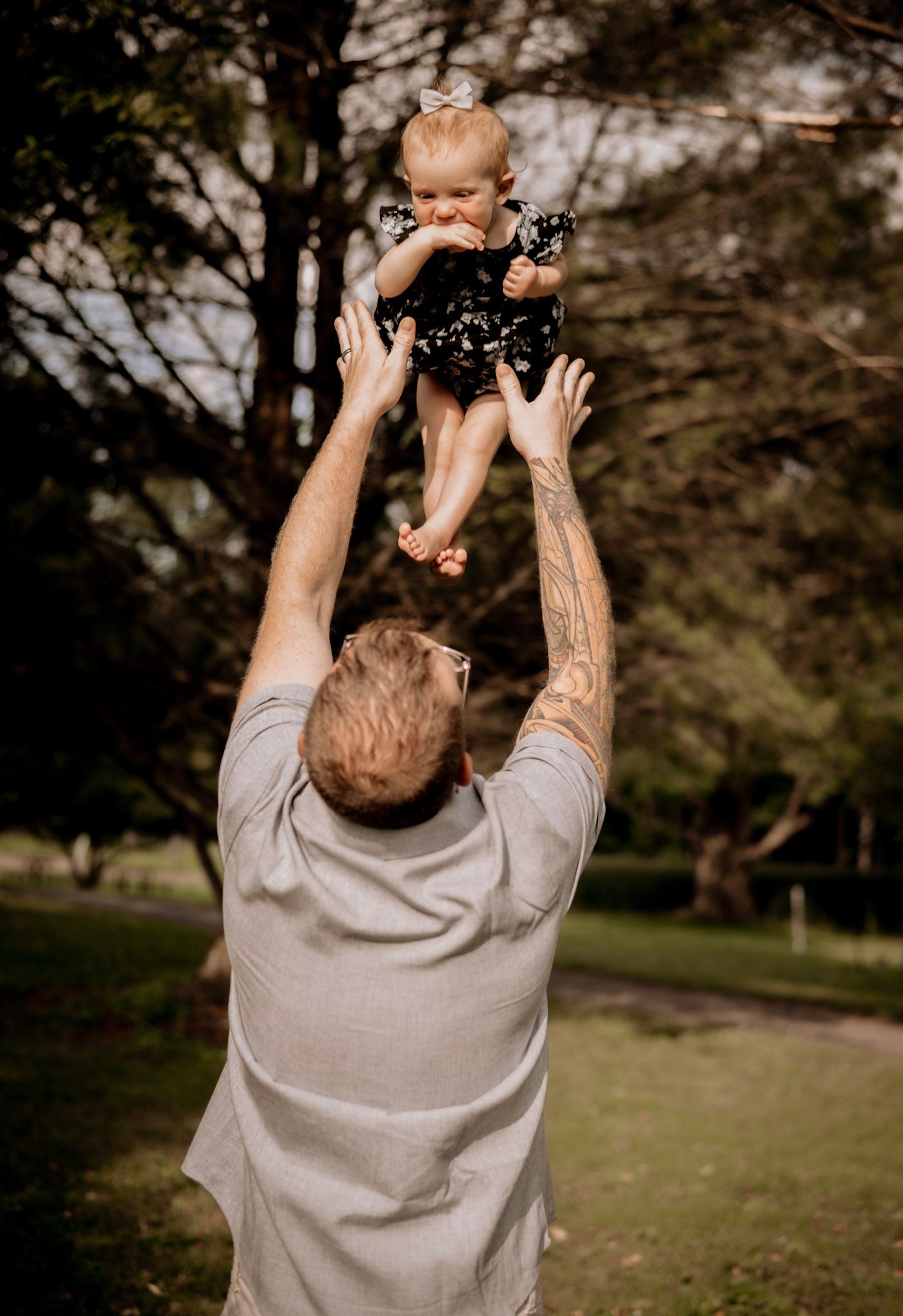 Four candid outdoor photos show a person in a gray sweatshirt lifting a small baby up into the air during a sunny day in a park.