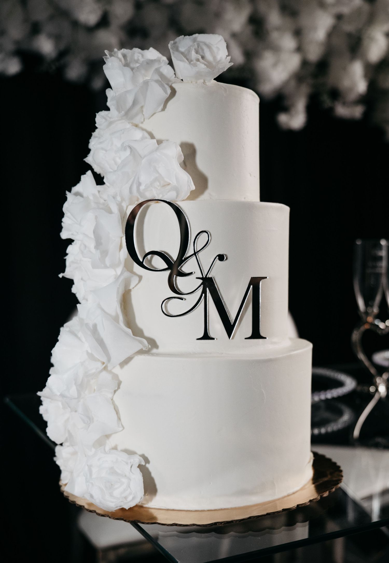 Elegant three-tier white wedding cake decorated with roses and monogram Q&M in black lettering.