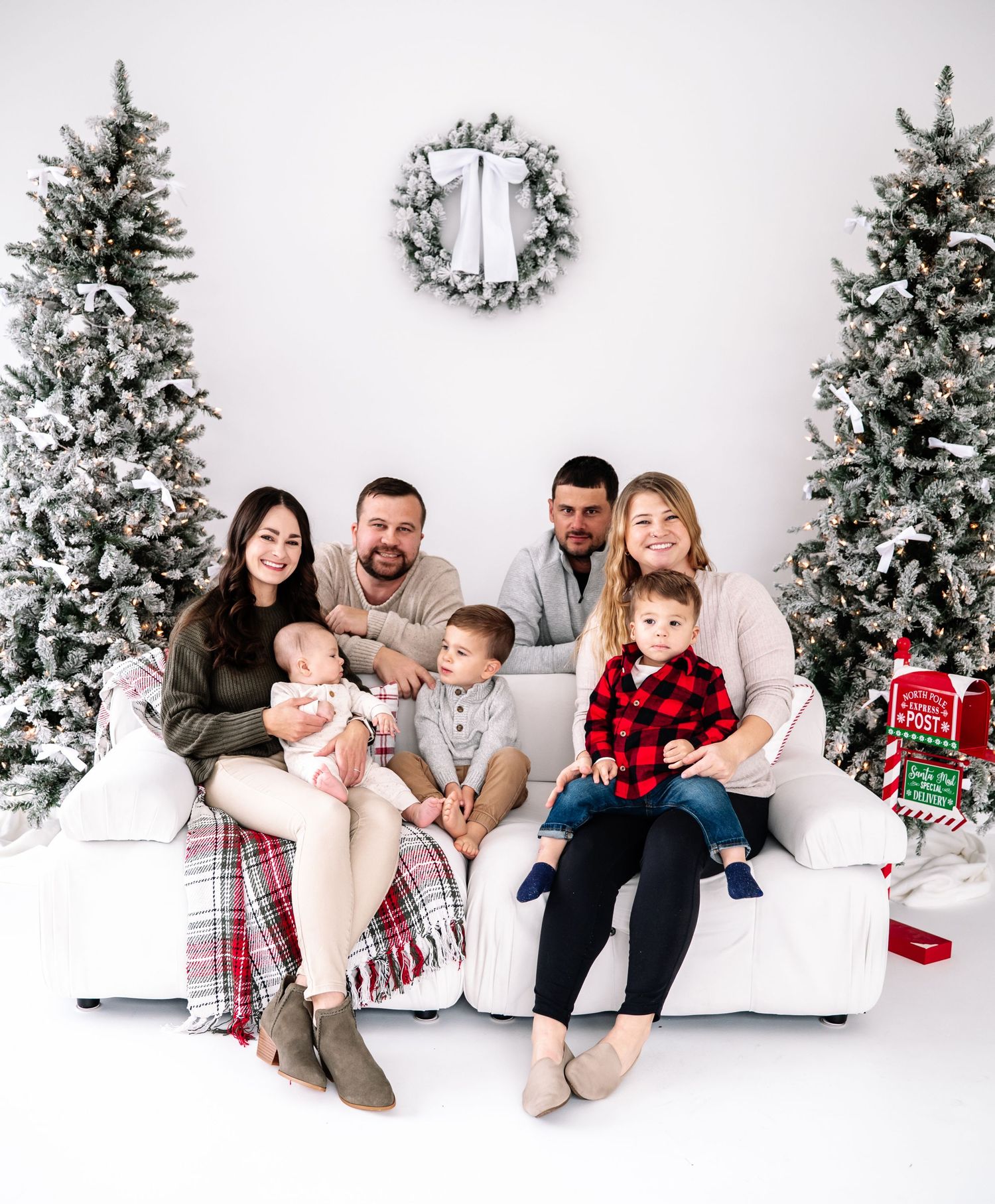 A family in coordinated winter outfits sits together against a white backdrop with snow-covered Christmas trees.