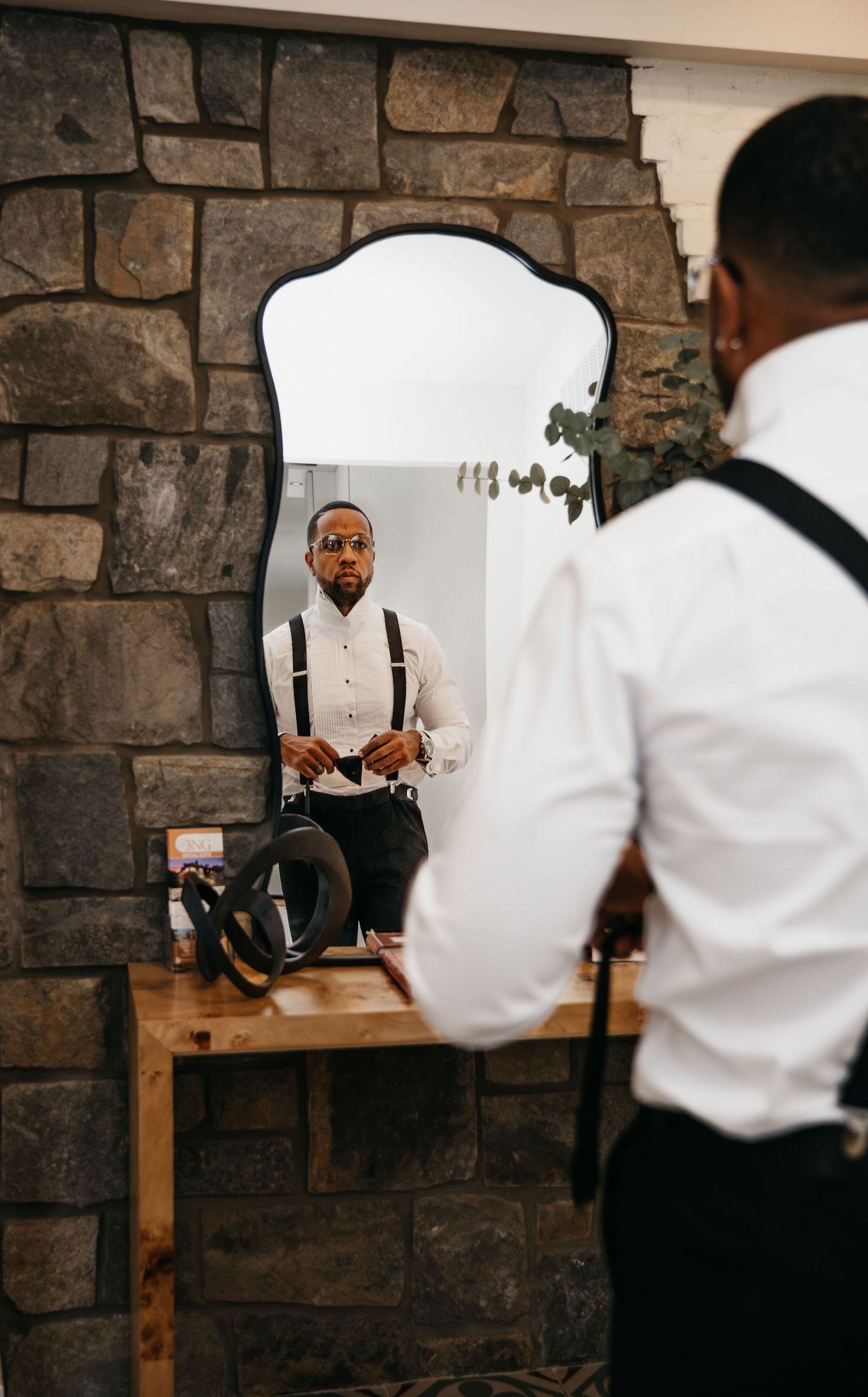 Reflection in stone wall mirror shows person in white dress shirt and suspenders getting ready.