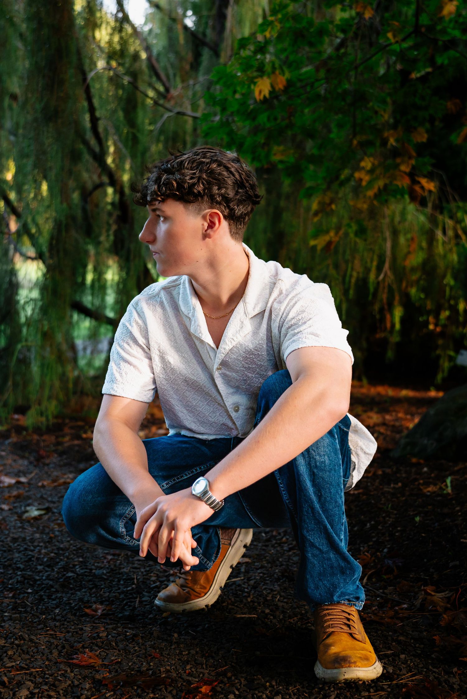 Person in white shirt and jeans crouches on dark ground in wooded area during autumn.