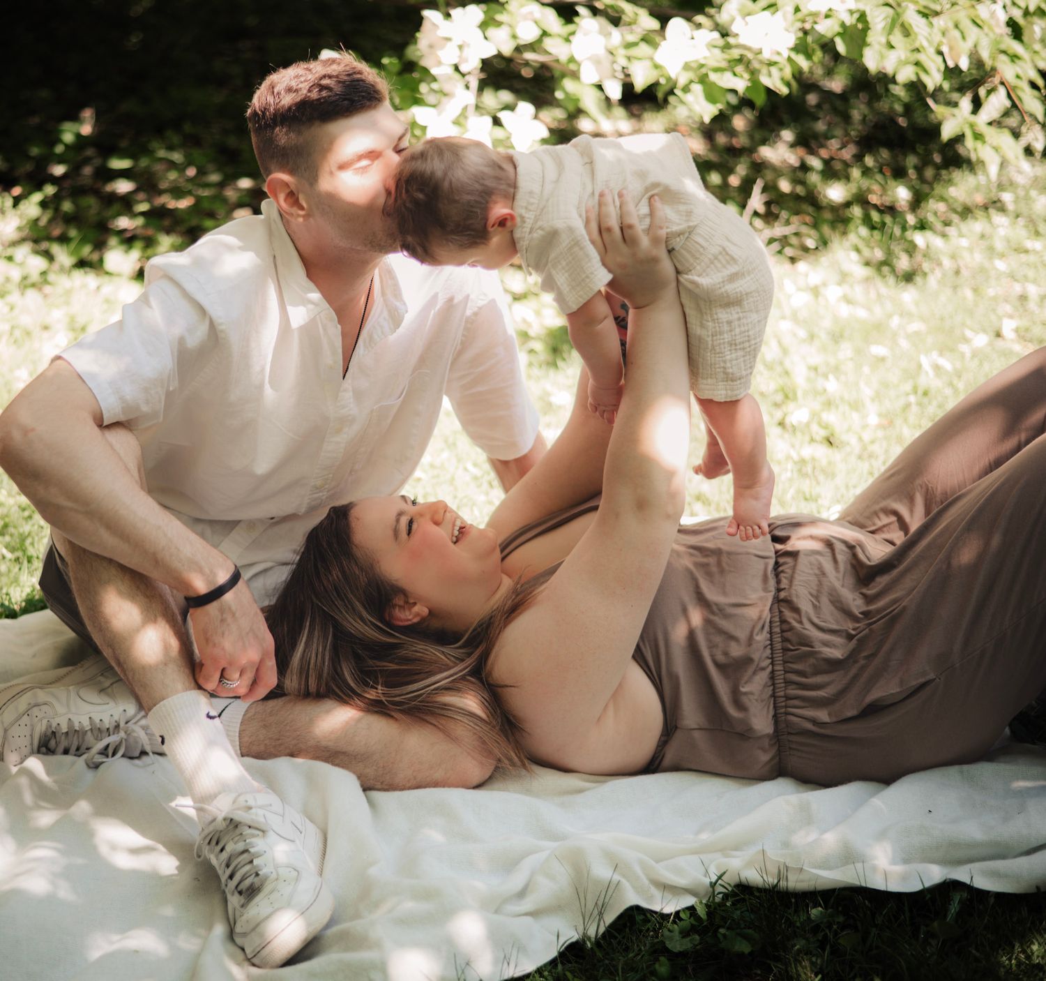 Candid family photo session outdoors on a white blanket in dappled sunlight.